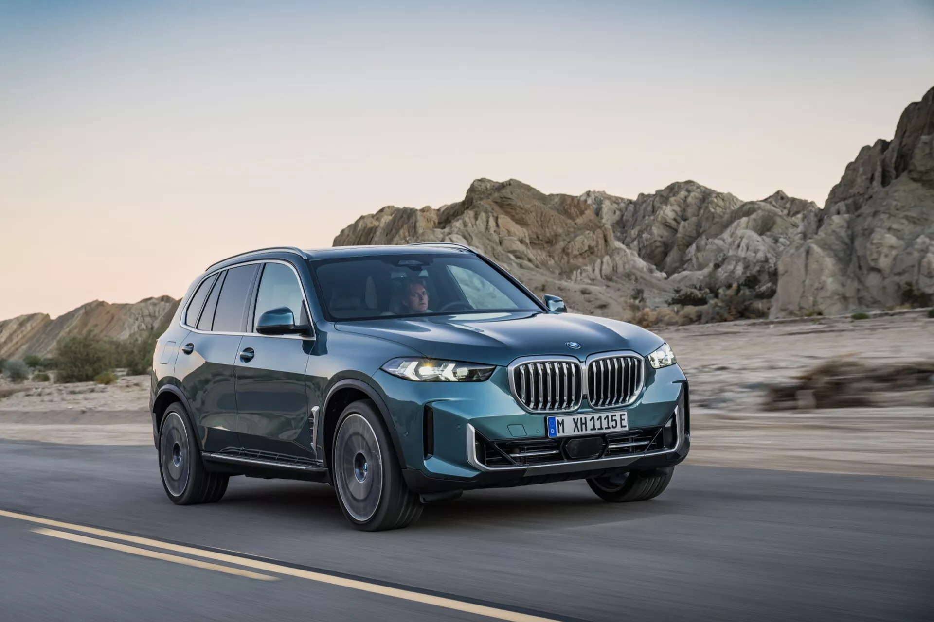 BMW X5 2023 (G05-F95-Facelift) i rörelse på en ökenväg, visar framgrillen, LED-strålkastare, flerspakshjul och blågrön färg från ett tre kvarts främre vy under sen eftermiddag