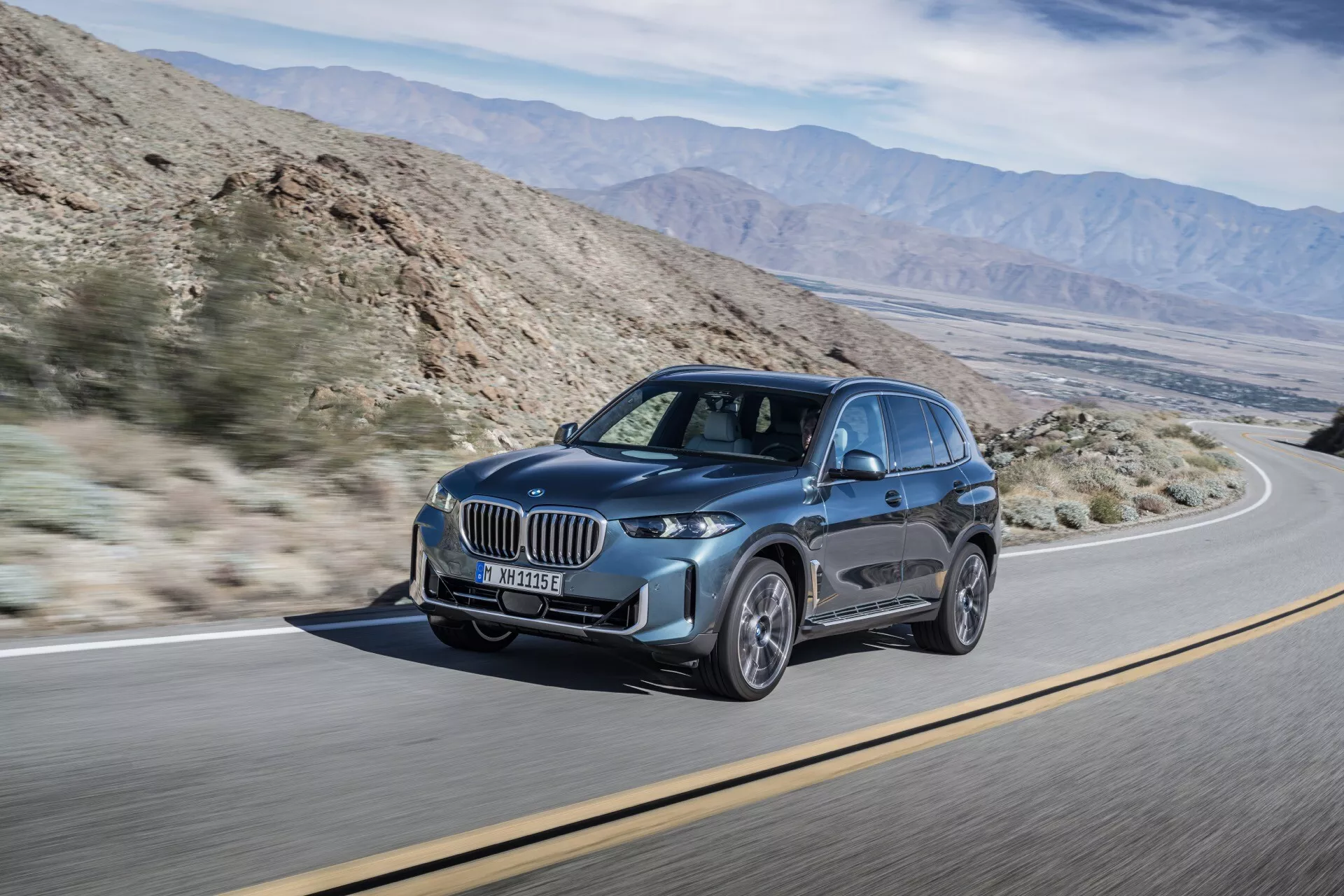 Lyxig SUV BMW X5 2023 (facelift G05-F95) i dynamisk rörelse, visar upp sin moderna design med en framträdande njurformad grill, slanka LED-strålkastare med dagljus, stora fler-ekrade legeringshjul, och skulpterade aerodynamiska karosslinjer mot en ökenliknande bakgrund med bergskedjor, framhävande funktioner som avancerat ljussystem och säkerhetsteknik.