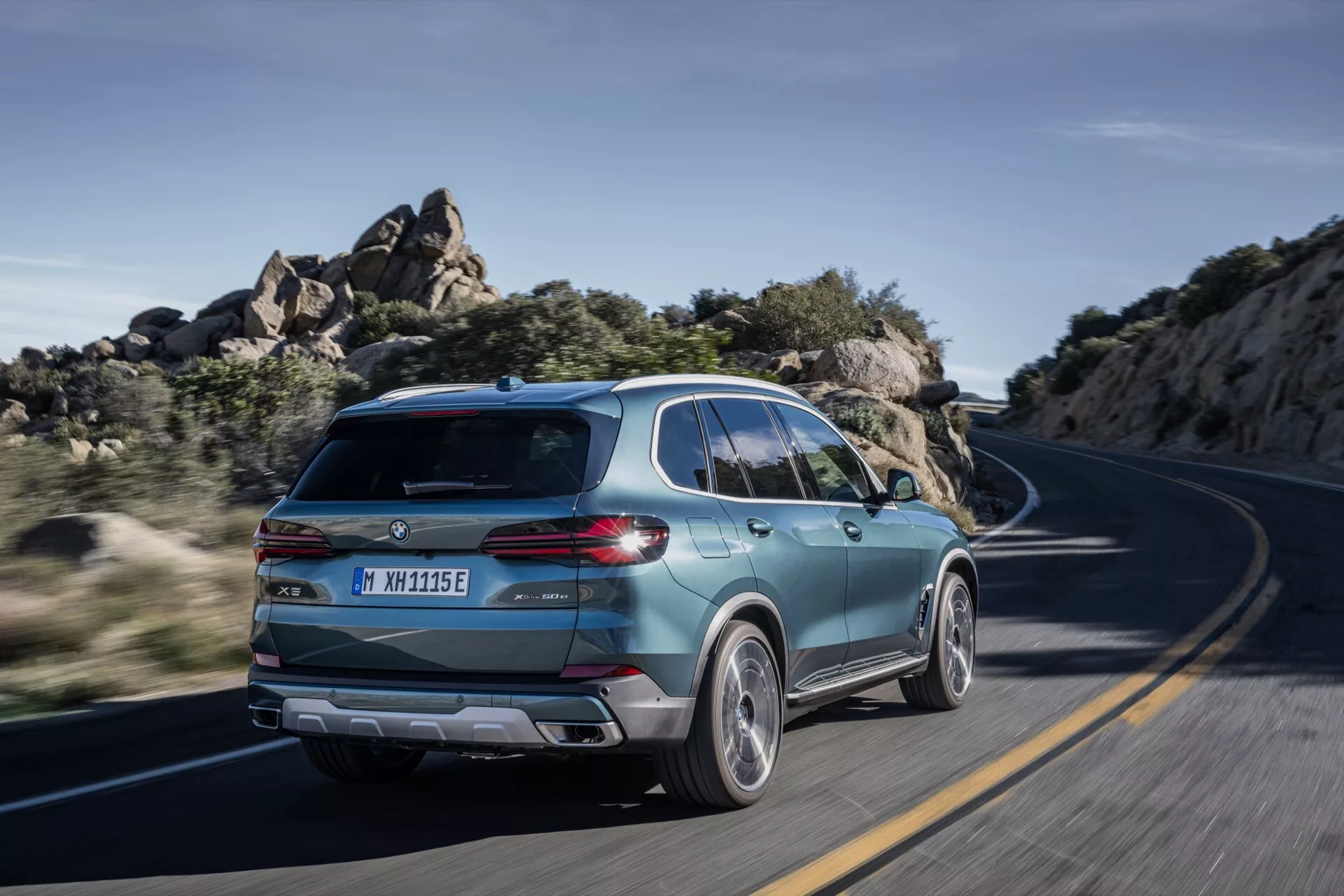 2023 BMW X5 (G05-F95 Facelift) i rörelse, fångad från en bakre trekvarts vy som framhäver dess sportiga SUV-design, moderna baklucka med snygga bakljus, tvåfärgade legeringshjul och tecken på elektrifiering på en slingrande väg under klart dagsljus