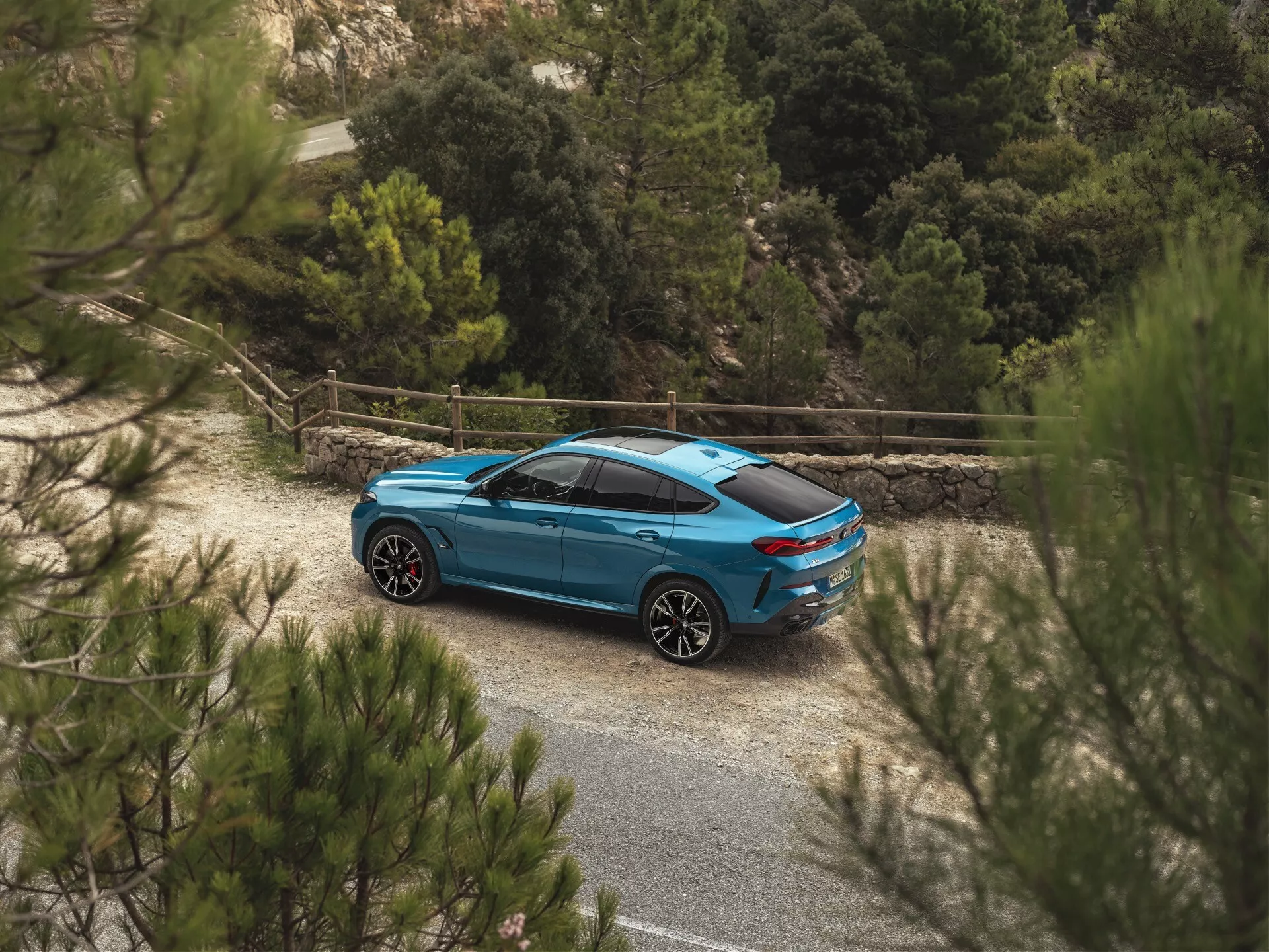 BMW X6 2023 (G06-F96-Facelift) i blått, fotograferad från ett högt höger bakifrån trekvarts perspektiv som framhäver dess sida och bakprofil, aerodynamisk design, anpassade hjul och integrerade teknikfunktioner mitt i en lantlig miljö med naturlig vegetation