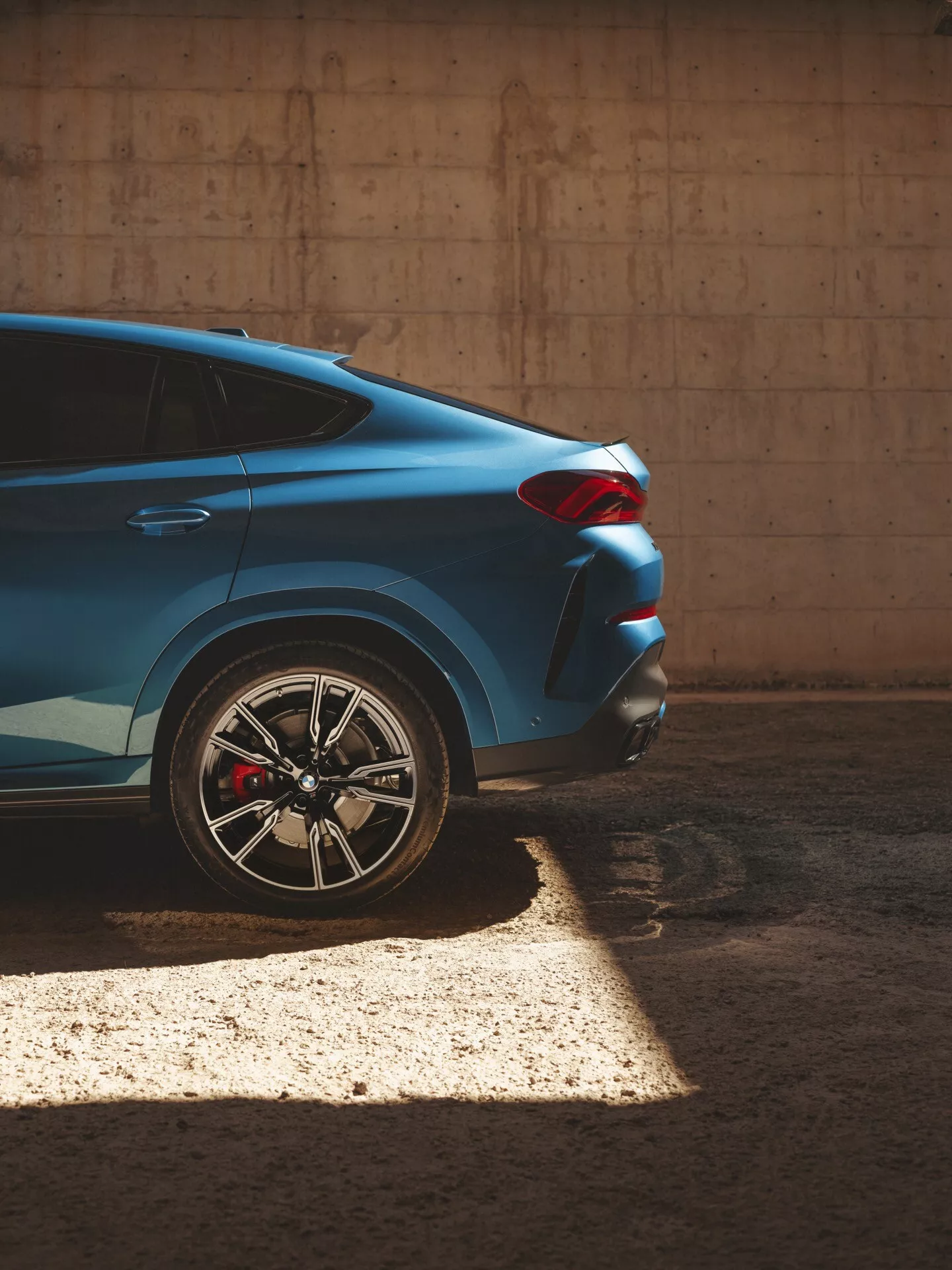 Blå BMW X6 2023 (G06-F96-Facelift) sedd från delvis sida och bakifrån som lyfter fram dess högra bakre kvart med fokus på det flerspakiga legeringshjulet, röda prestanda bromsoket och det skarpa, vinklade designen på bakljuset, fångat i ett skuggigt ljus som skapar en elegant dynamisk kontrast mot en betongbakgrund.