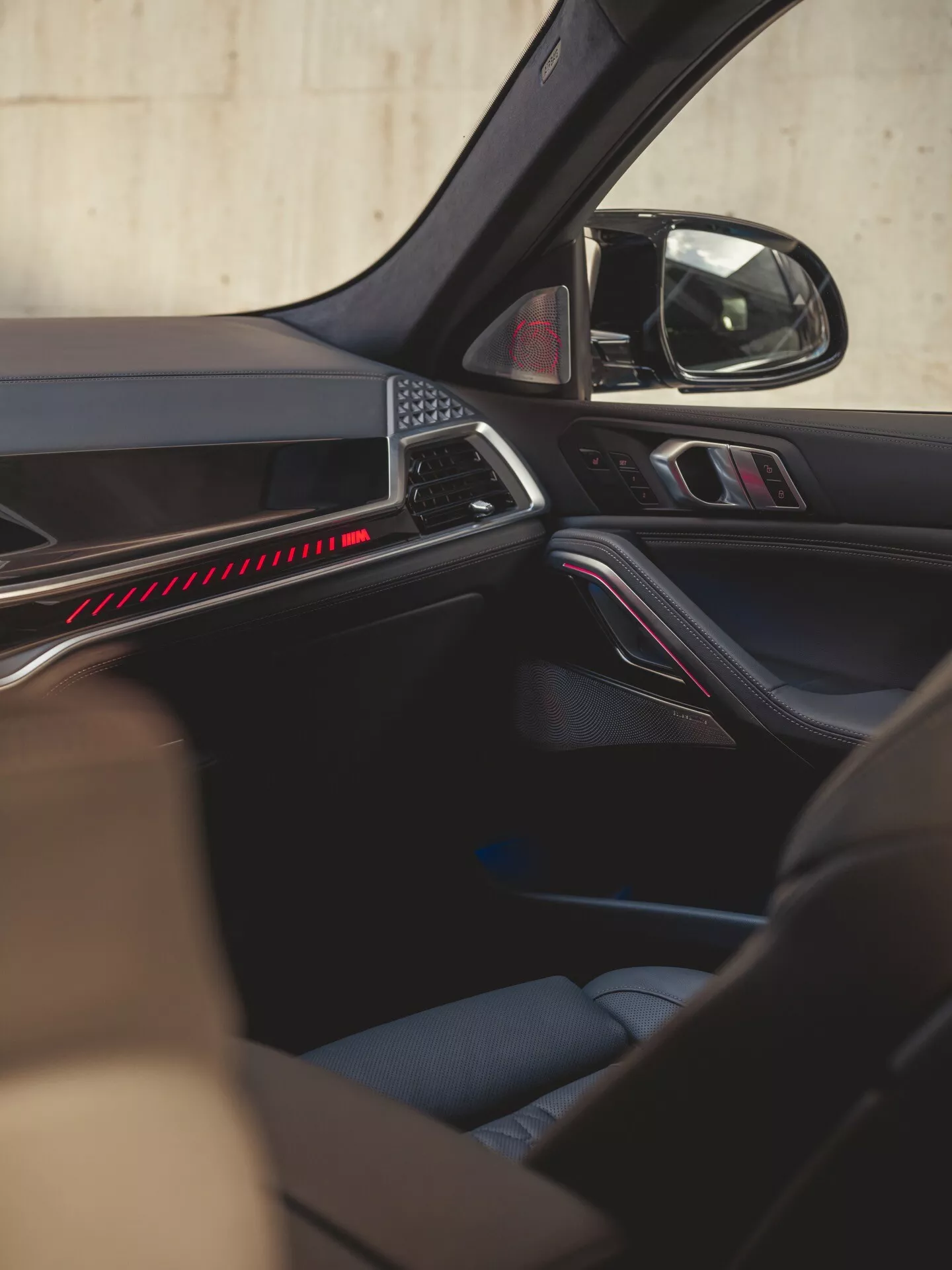Interiörvy av BMW X6 2023 (G06-F96 Facelift) med fokus på detaljer på passagerarsidan dörren, med ergonomisk panel design med röd accentbelysning, premium material finish och kontroller för elektriska fönster och speglar, kompletterat med en högkvalitativ ljudsystemhögtalare, och säkerhetsorienterad design med synligt säkerhetsbälte, illustrerar komfort, bekvämlighet, ljud, belysning och säkerhetsteknologier.