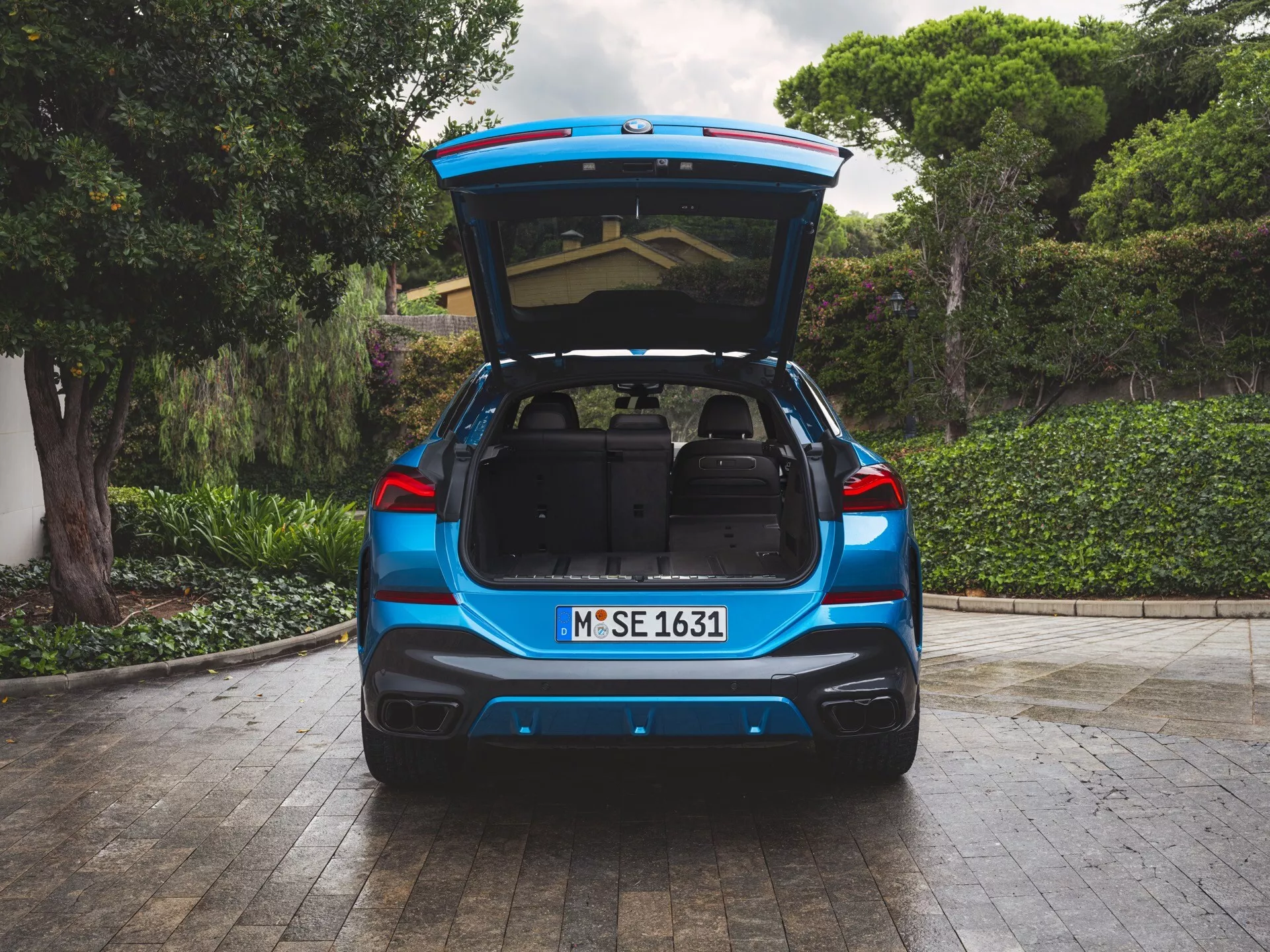 Baksida vy av blå BMW X6 2023 (G06-F96-Facelift) med öppen bagagelucka och bagage, som visar rymligt lastutrymme, parkerad på en uppfart med naturlig vegetation i bakgrunden, med München registrering, parkeringssensorer och dubbelt avgassystem