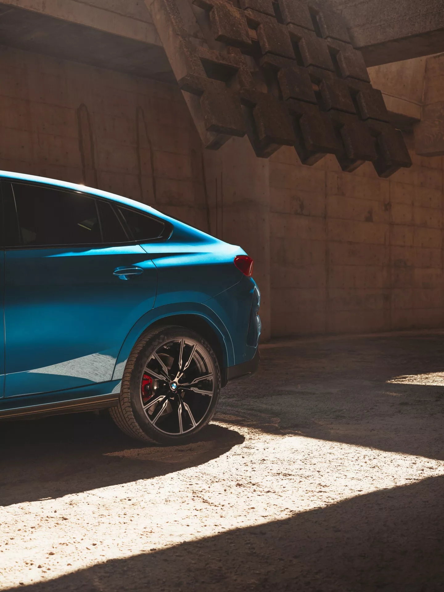 Blå bil BMW X6 2023 (G06/F96 facelift) i solljus, sedd från ett tre fjärdedels bakre perspektiv som betonar den bakre fjärdedelen med sportig fälg och röd bromsok.