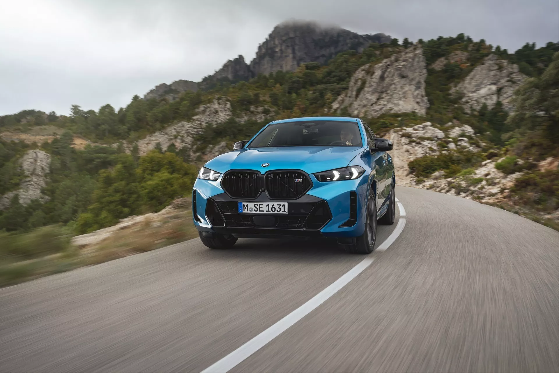BMW X6 2023 (G06-F96-facelift) i livligt blått, visar modern lyxprestanda på en slingrande bergsväg med dynamisk rörelseoskärpa, avancerade LED-strålkastare, eleganta designelement och sportiga hjul, mot en bakgrund av robust bergsterräng.