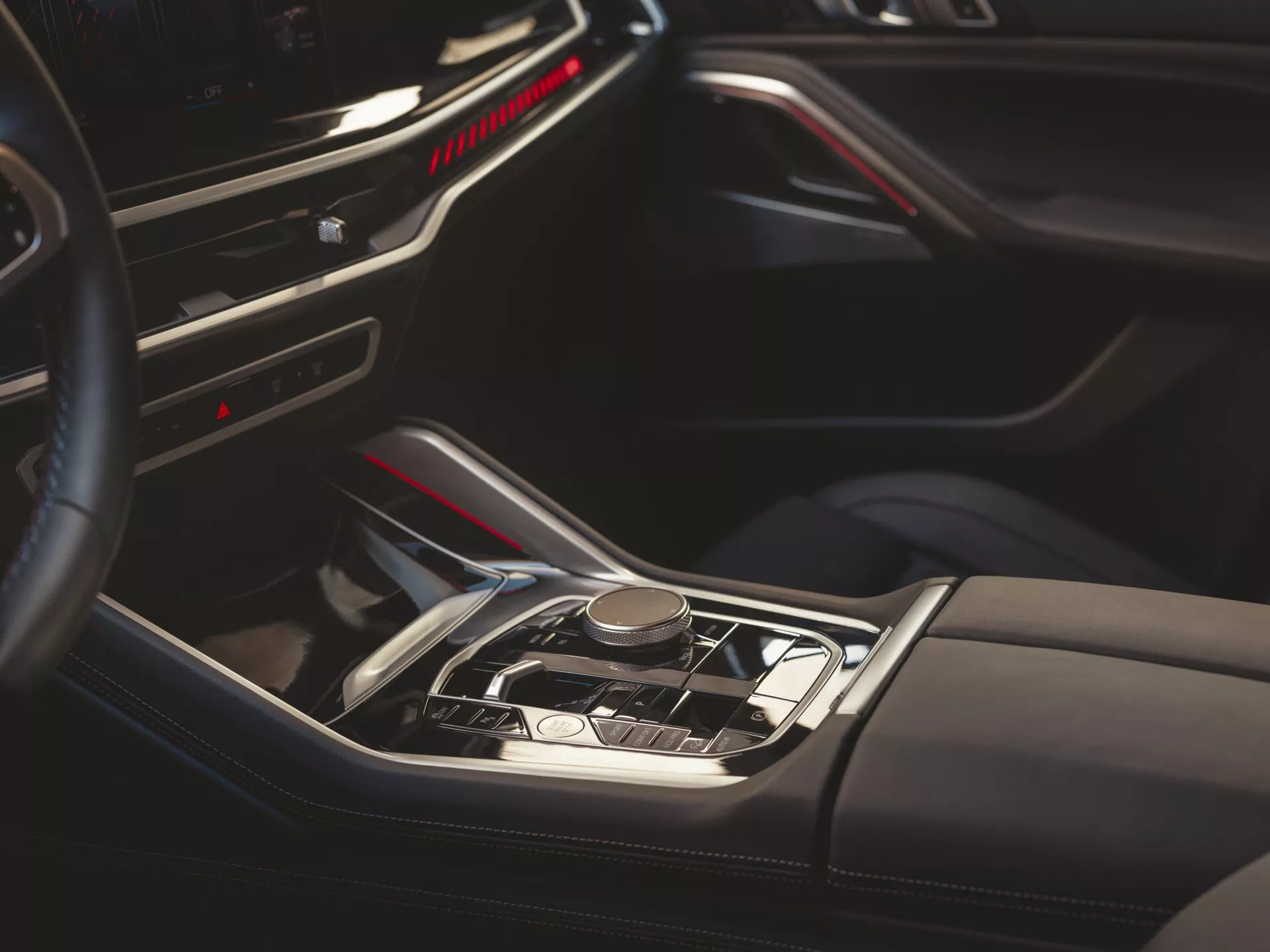 Närbild av interiören i BMW X6 2023 (G06-F96-Facelift) som visar den automatiska växelspaken, den lyxiga mittkonsolen med träliknande dekor, metalliska accentdetaljer, röd ambient belysning, läderdetaljer och delvis vy av instrumentbrädan och förarens dörr