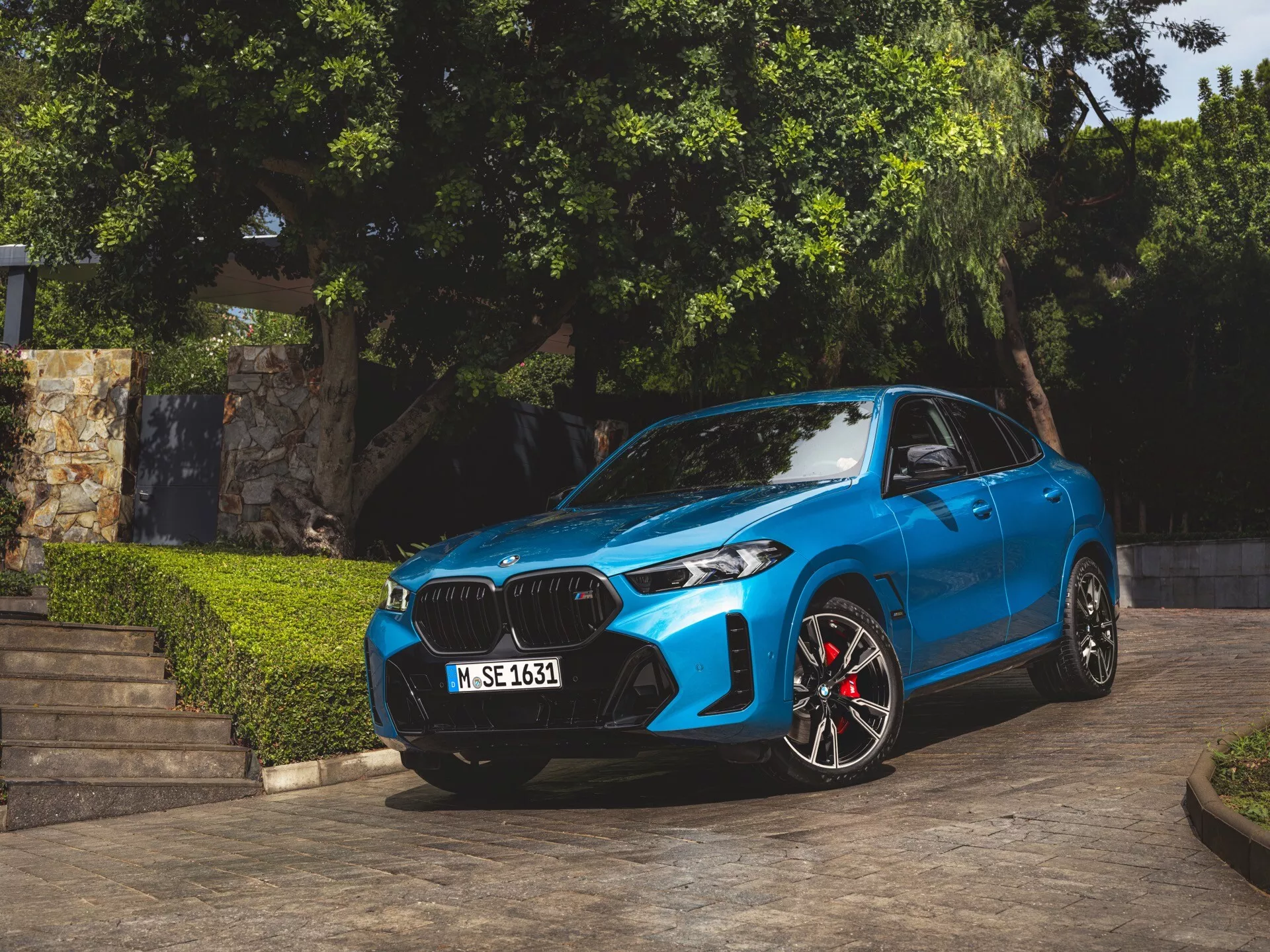2023 BMW X6 (G06-F96-Facelift) i blått, parkerad i vinkel som visar dess främre fasad, njurformad grill, LED- eller laserstrålkastare, sportiga legeringshjul med röda bromsok, konturerade huvar och aerodynamiska sidbackspeglar med integrerade blinkers, fångad i naturligt dagsljus med grönska och arkitektoniska steg i bakgrunden