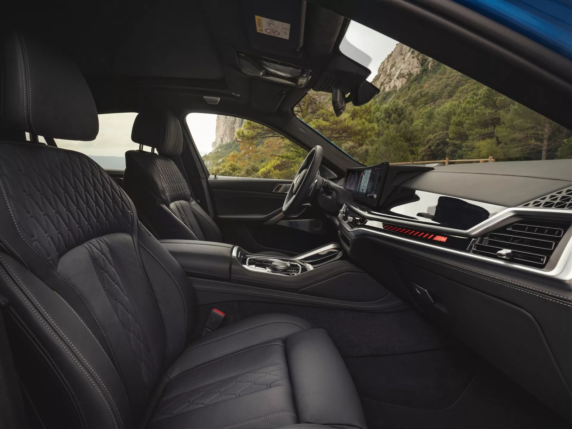 Interiörvy av BMW X6 2023 (G06-F96 Facelift) från baksätet som visar fram instrumentbrädan, framsäten i premiumläder med quiltad design, mittarmstöd och avancerat touchscreen infotainmentsystem, omgivet av en modern, minimalistisk estetik som indikerar lyx och banbrytande fordonteknologi