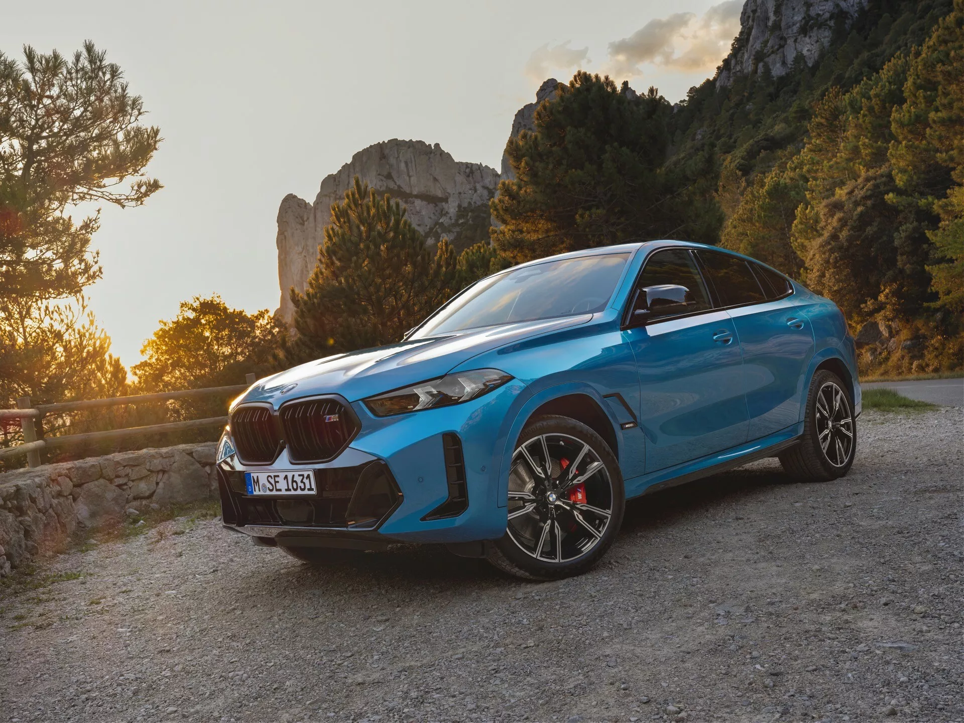 BMW X6 2023 (G06-F96-Facelift) i blått, med en sportig SUV-design fångad under gyllene timmen med en framifrån tre fjärdedels vy som visar den distinkta stora njurgrillen, slanka LED-strålkastare och prestandaorienterad frontstötfångare med stora luftintag, mot en bakgrund av en lugn bergsväg.