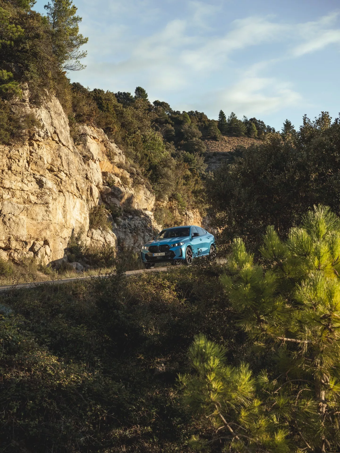 BMW X6 2023 (facelift G06-F96) i tre fjärdedels framvy på en slingrande bergsväg omgiven av skog, visar terränggående förmåga och modern design