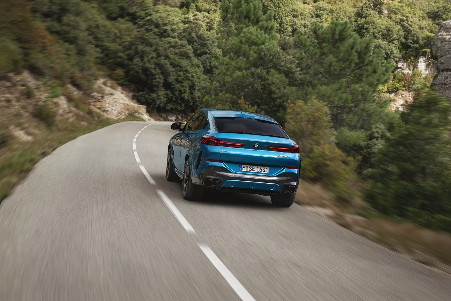 Dynamisk bakvy av en blå BMW X6 2023 (G06-F96-Facelift) med sportig design och moderna bakljus på en slingrande väg mitt i ett naturlandskap