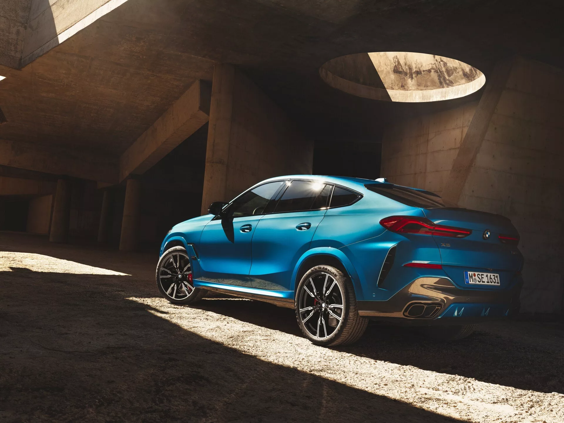 BMW X6 2023 (G06 F96 facelift) i blått, fångad från en trekvarts vinkel bakifrån som framhäver fordonets designlinjer, silhuett, formen på bakfönstret, bakhjulsbågarna och bakljusen, parkerad på betong under kontrasterande solljus.