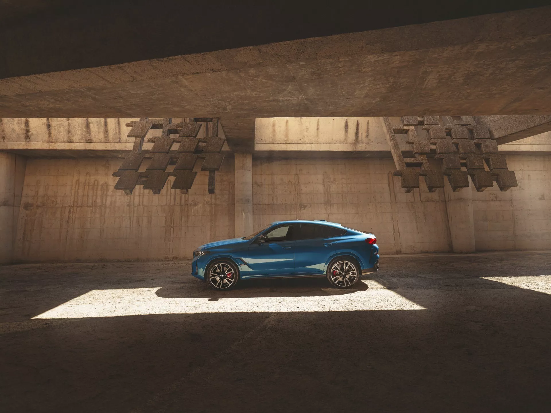 2023 BMW X6 (G06-F96 Facelift) kupéstil bil i glänsande blått, framhävd av solljus genom en öppning ovan, fångar dess slanka sidoprofil, aerodynamiska linjer och lyxiga funktioner som legeringshjul och soltak