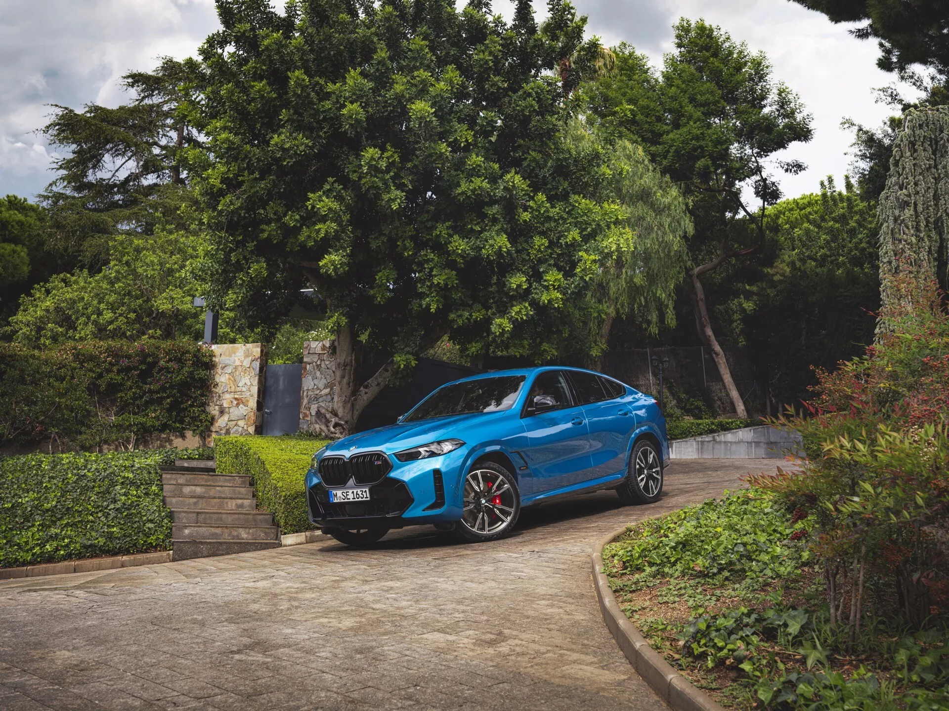 BMW X6 2023 (G06-F96-Facelift) i blått, tre fjärdedels framifrån vy, parkerad på en uppfart omgiven av grönska, visar frontstötfångare, grill, huva, vindruta, förarsidans profil, LED-strålkastare, sportiga hjul med röda bromsok och aerodynamisk design