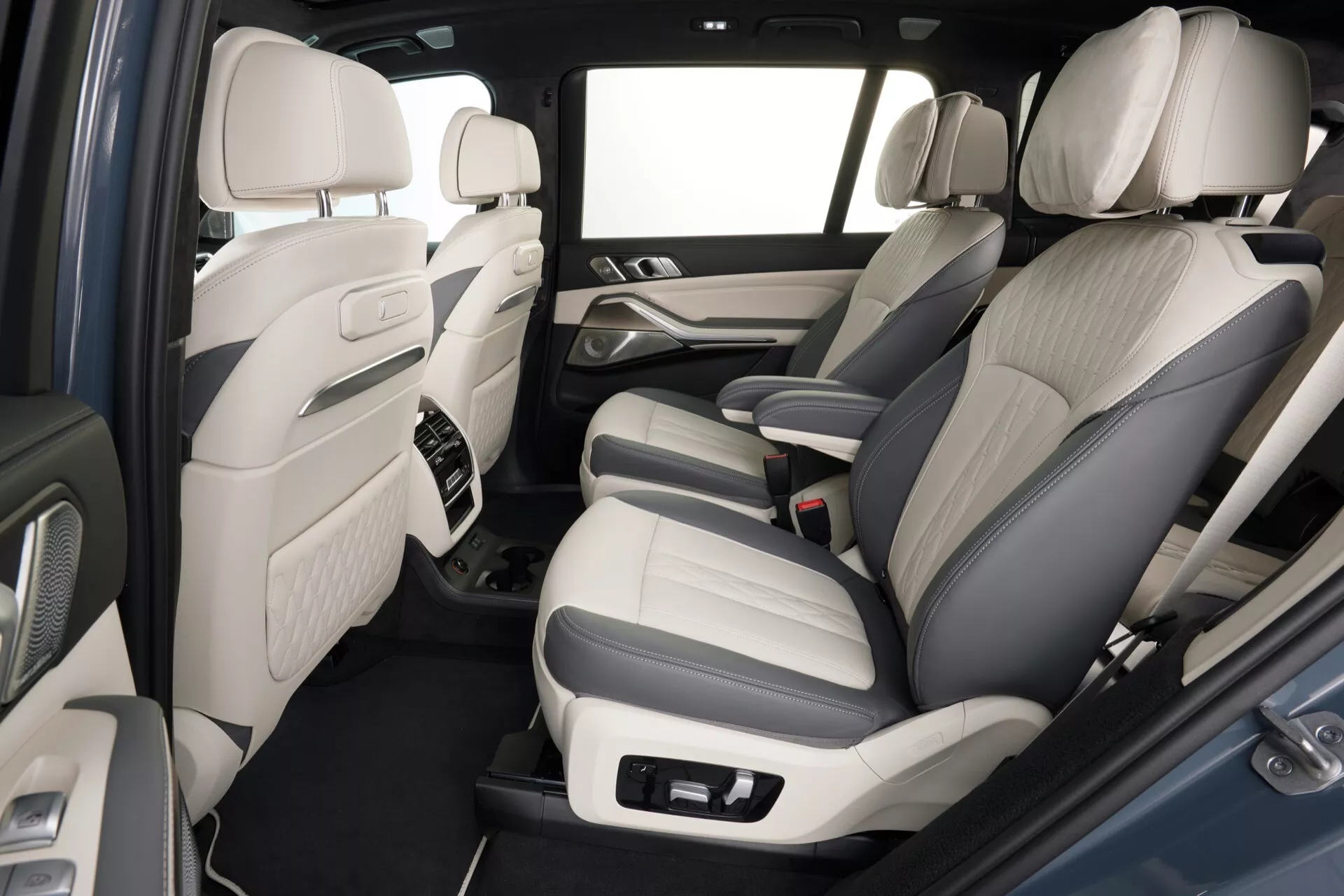 Interiörvy av BMW X7 (G07 Facelift) 2022 med fokus på det bakre passagerarutrymmet, med tvåtonigt läderklädsel, avancerade ljudsystemskontroller på dörrpanelerna, mittkonsol med högkvalitativa materialaccent, mattat golv, gott om benutrymme och en ljus interiörfärgschemat för en rymlig kabin känsla