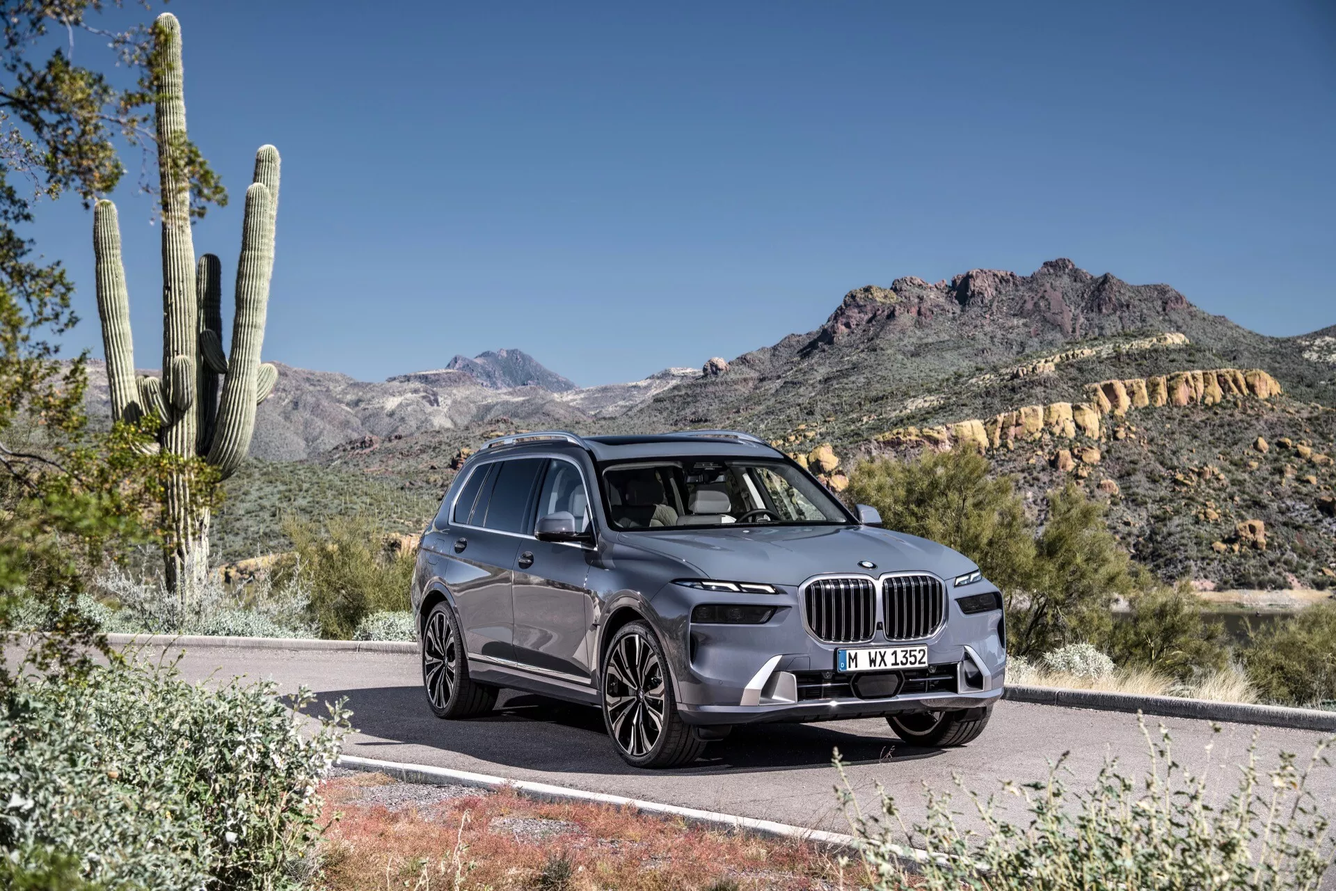 Trekvarts framsida av BMW X7 2022 (G07-Facelift) på en asfalterad väg, som visar dess stora njurformade grill, vinklade LED-strålkastare, skulpterad huva och förarsidans profil mot en öken- och bergsbakgrund i dagsljus.
