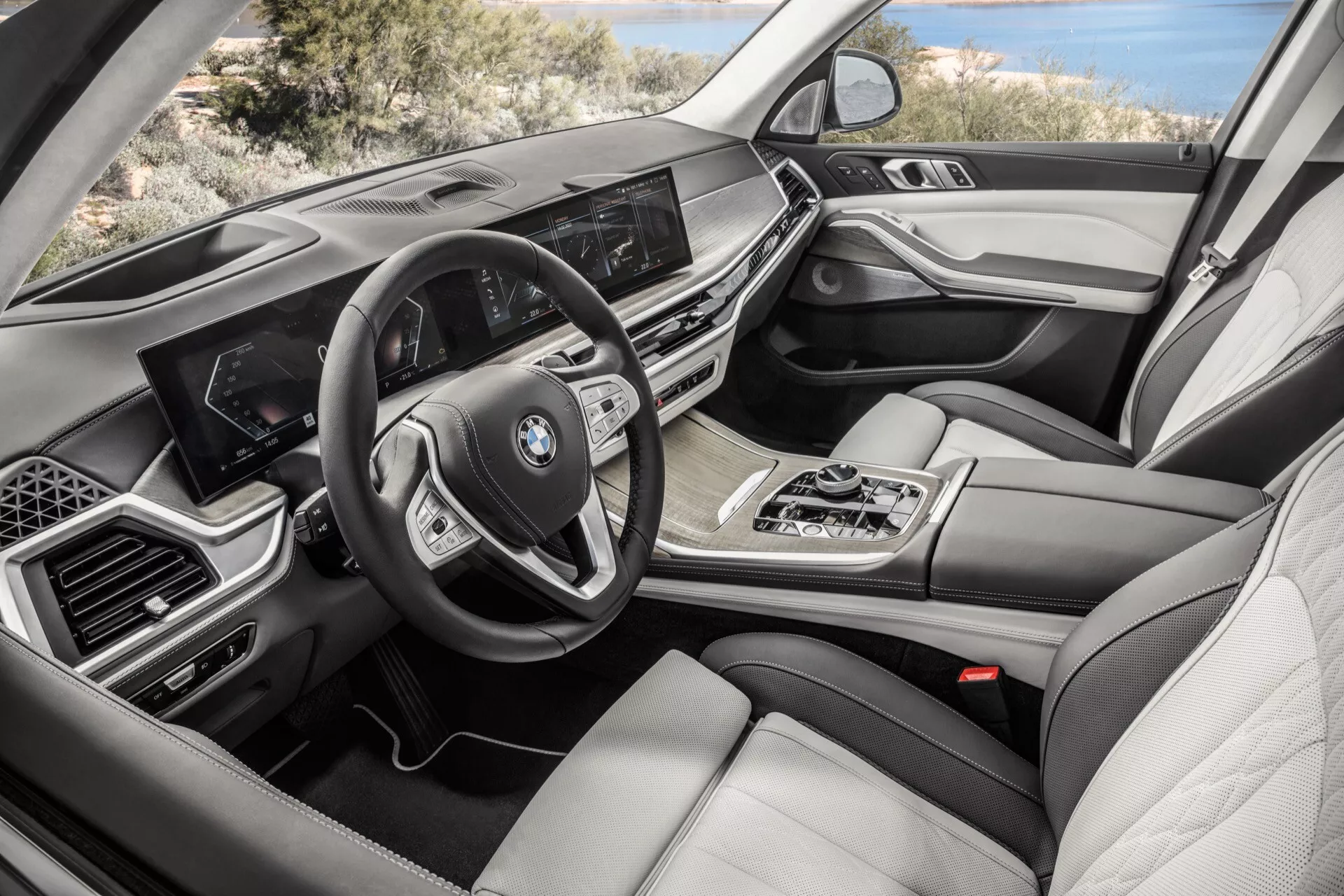 Interiörvy av en BMW X7 2022 (G07-Facelift) från passagerarsidan som framhäver instrumentbrädan med infotainmentskärm, multifunktionsratt, automatväxelspak, iDrive multimediekontroll, dubbelzons klimatkontroll, lyxiga lädersäten, trä och metallinlägg, samt digital instrumentpanel.