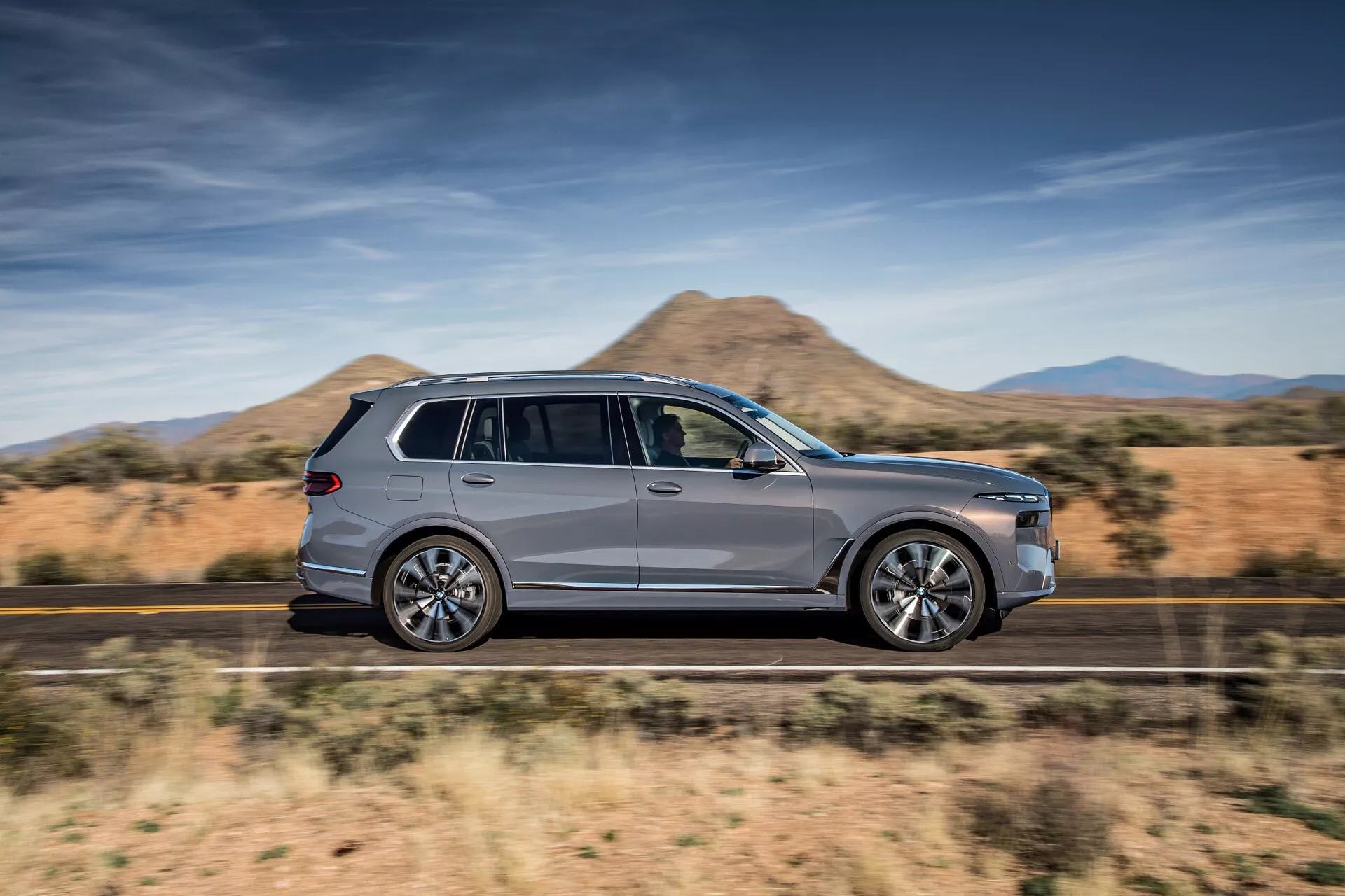 BMW X7 (G07 facelift) 2022 i rörelse, visar upp sin aerodynamiska design och lyxiga utseende från en sidoprofilvy på en ökenväg, med fokus på SUV:ens slanka siluett, metallicfärg, kromdetaljer, stora legeringshjul och funktioner hos taklastsystemet