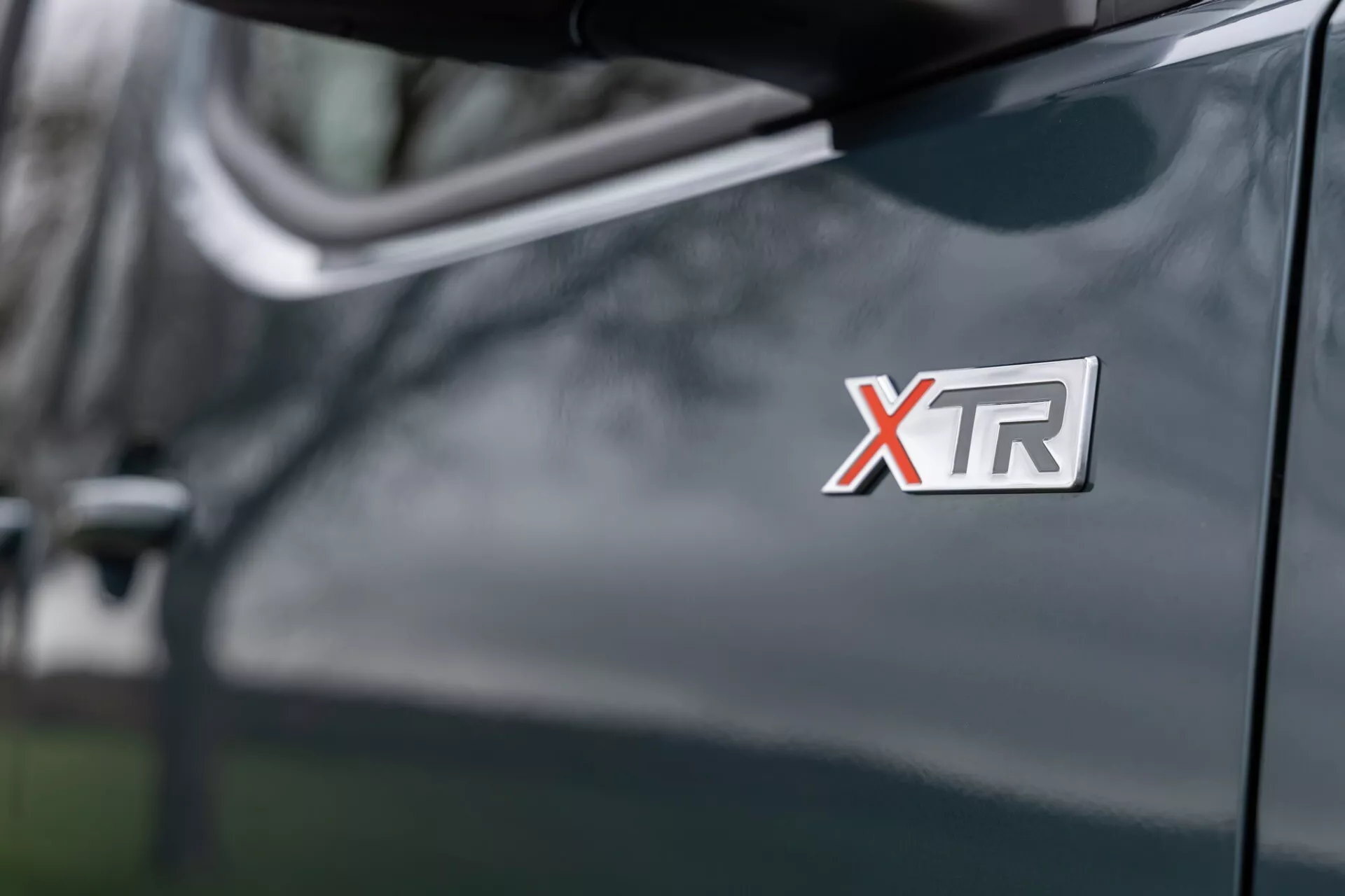 Närbild av Citroen Berlingo 2024 XTR-märket på framkanten, med en glansig mörk nyanserad finish med en sidospegel och suddiga utomhusreflektioner i bakgrunden.