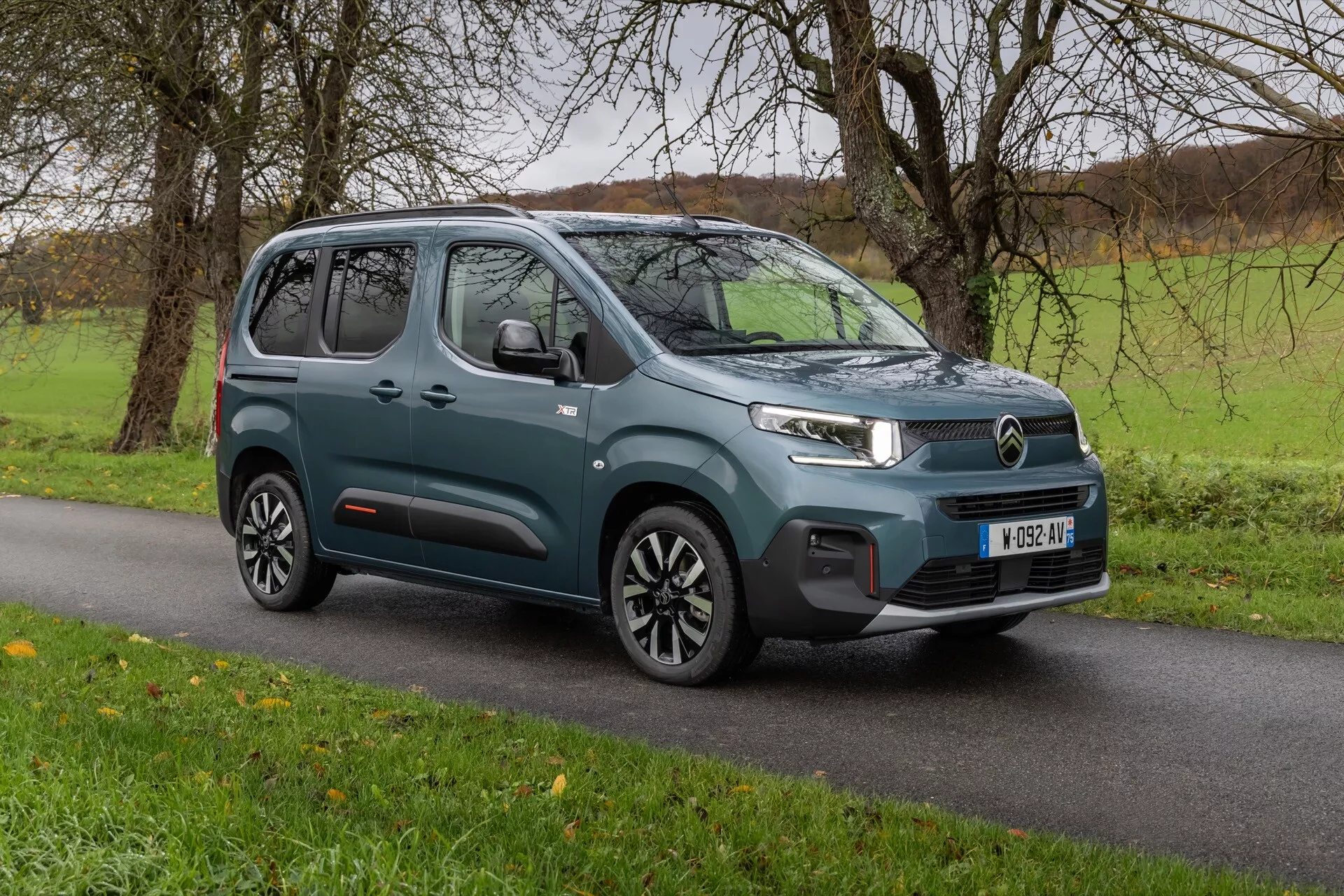 Facelift för 3:e generationens Citroen Berlingo 2024, modern kompakt MPV på asfalt med gräs i bakgrunden som visar en trekvarts främre vy, framhäver LED-strålkastare, märkesspecifik frontgrill, stötfångare med parkeringssensorer och snygga sidokonturlinjer med flerspakiga aluminiumfälgar i tvåfärgsschema.