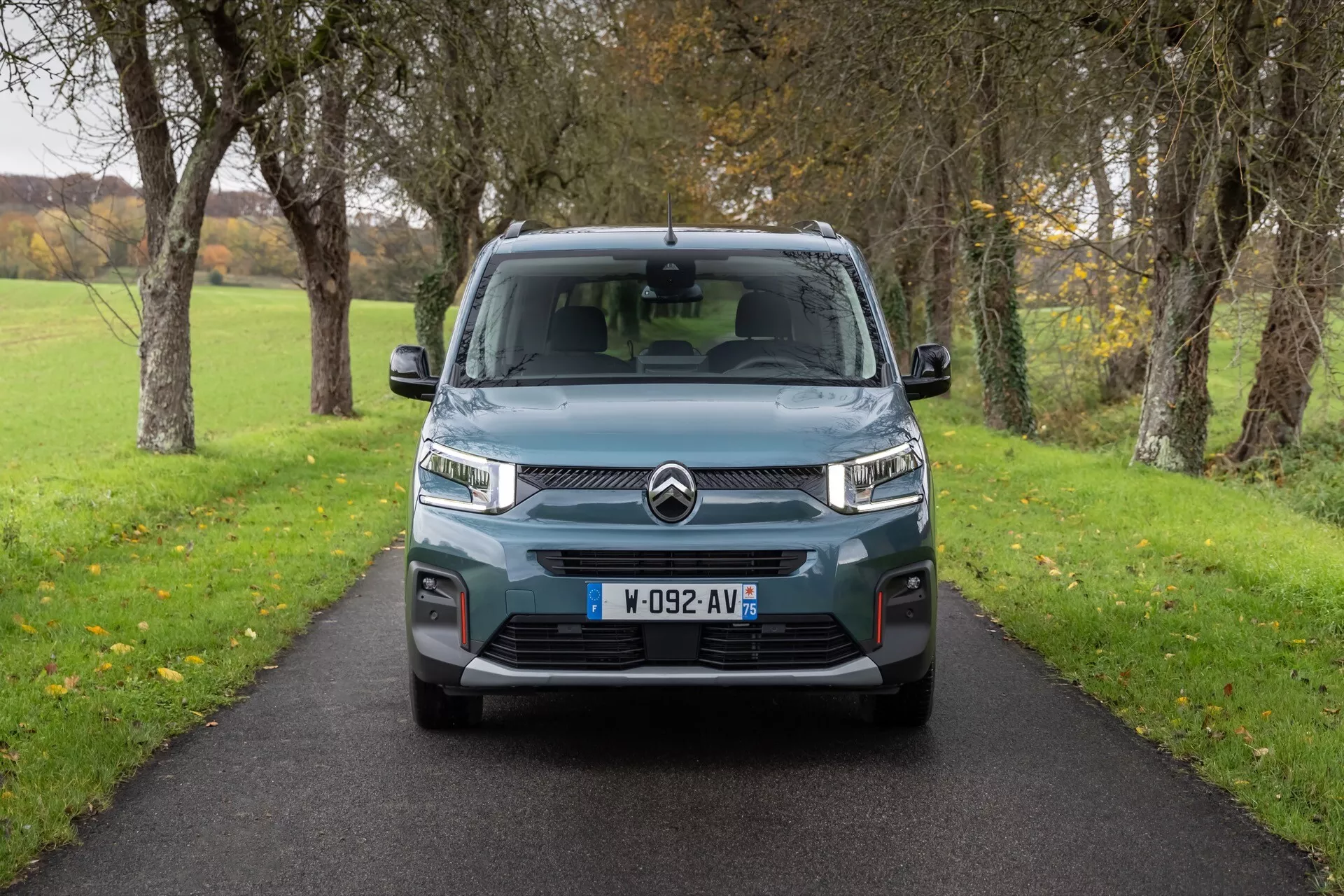 Citroën Berlingo 3:e generationens faceliftmodell 2024 fångad framifrån på en smal väg, med LED-dagsljus, invecklad grilldesign med dubbel-chevron-logotyp och parkeringssensorer