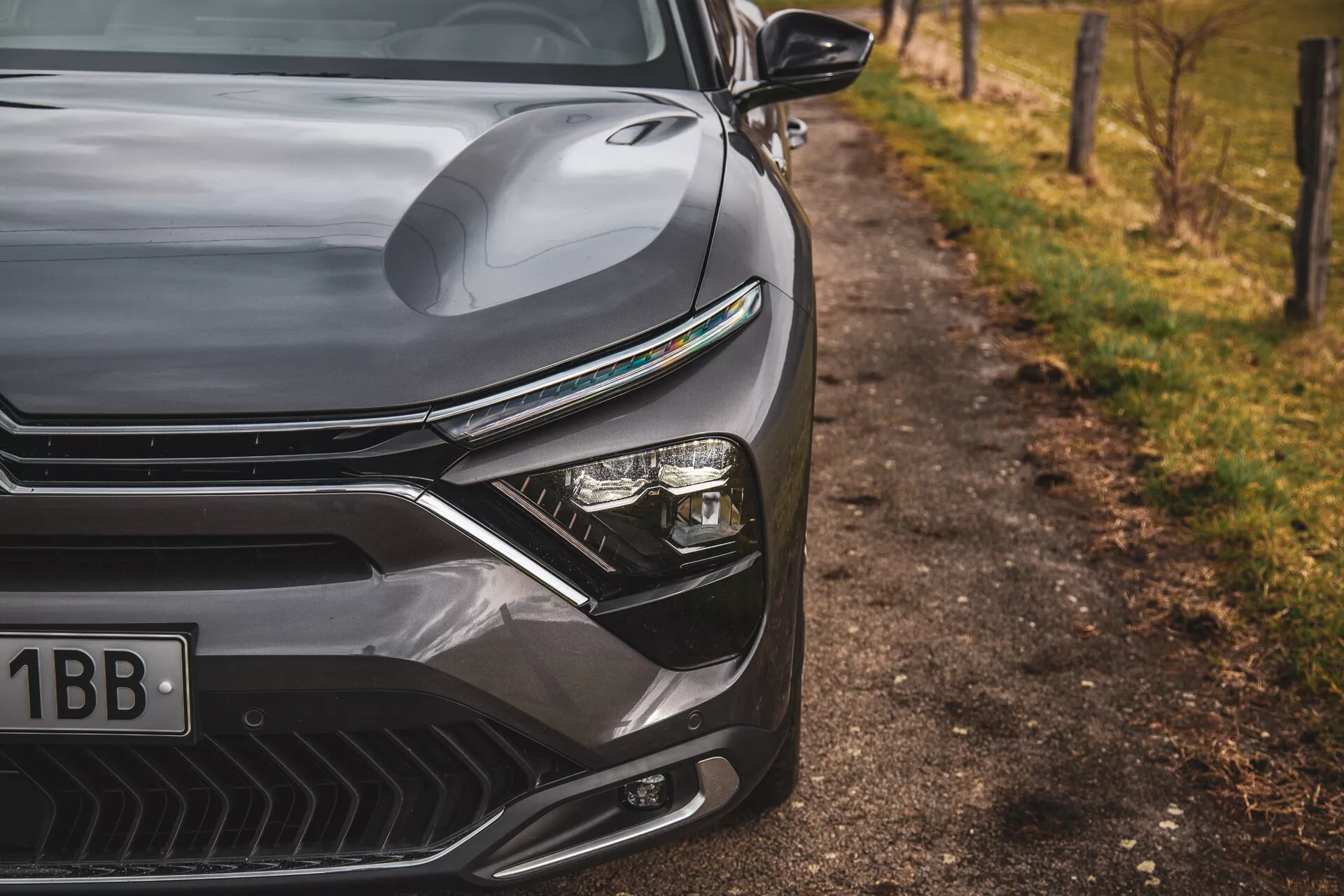 Närbild av Citroen C5-X 2022 som visar den eleganta designen på framsidan till vänster, inklusive det konturerade motorhuven, uttalad grill med horisontella linjer, avancerat LED-strålkastarsystem och aerodynamiskt formad frontstötfångare med luftintag, mot en suddig naturlig bakgrund vid vägkanten.