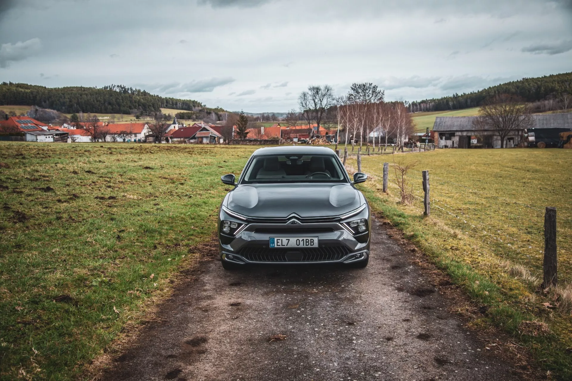 Frontvy av en Citroën C5-X 2022 på en smal stig i ett gräsfält under en molntäckt himmel, visar dess eleganta design, intrikata grill med emblem, vinklade LED-strålkastarenheter, skulpterade huvar, och ytterligare framlyselement, registrerad i Tjeckien.