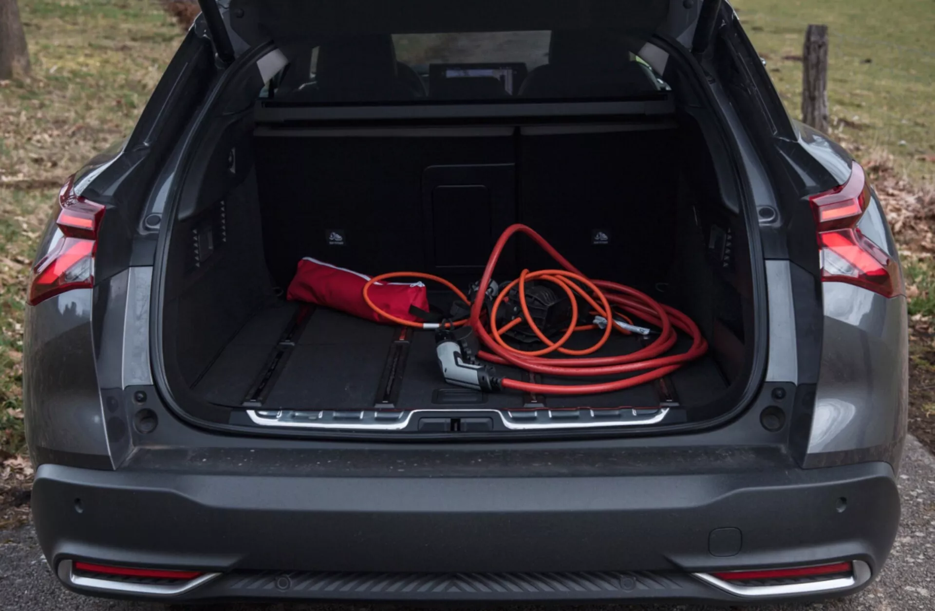 Vy av Citroen C5-X 2022 bakre bagageutrymmet med öppet fack som visar ett rymligt lastutrymme, röd-svart väska, orange förlängningssladd, extra förvaringsfack, skyddslist och parkeringssensorer i naturligt utomhusljus
