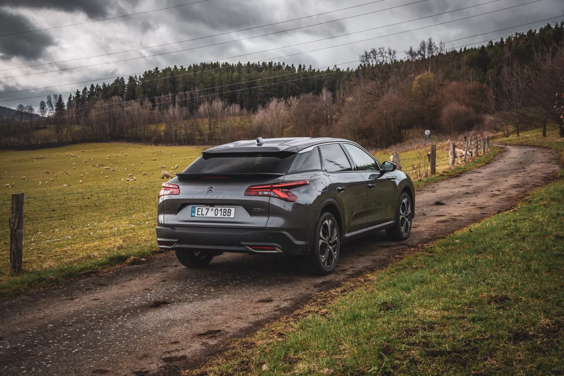 2022 Citroen C5-X modernt halvkombi parkerad vid en grusväg på landsbygden, vy från bakre tre kvarts vinkel med aerodynamisk design inklusive spoiler och hajfena-antenn, smala horisontella bakljus och stora flerspakade hjul, antyder prestanda och lyx i en icke-stads inställning.