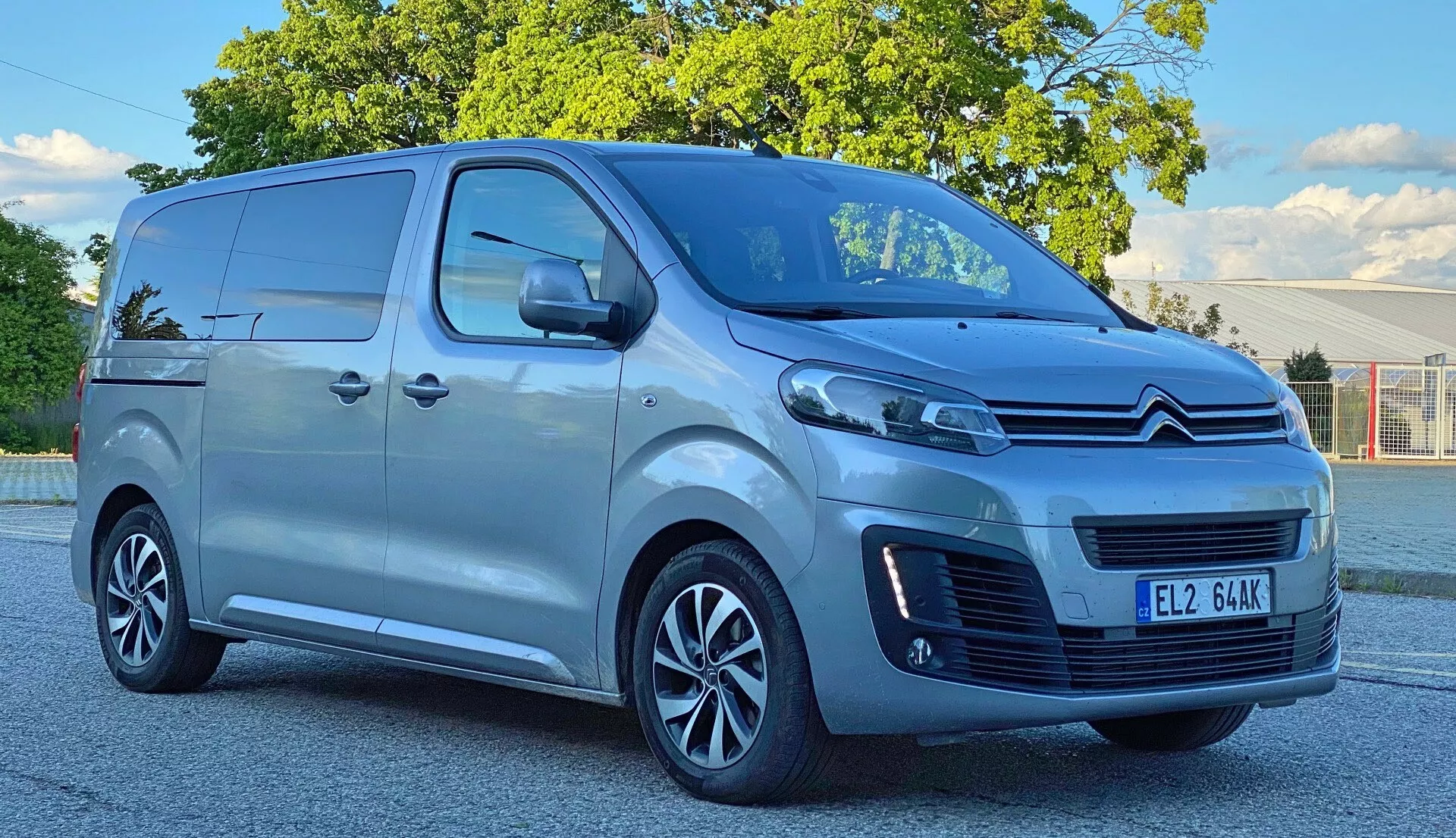 Låg vinkelbild av en ljusfärgad Citroen Spacetourer skåpbil från 2016, med fokus på dess höjd och storlek. Skåpbilen har ett krombelagt galler med Citroens dubbel-vinkel emblem, en bred luftintagssektion med horisontella lameller, eleganta projektionsstrålkastare och svarta konturerade dimljushålor. Sidosikten visar mångekrade lättmetallfälgar, skjutdörrar, stora sidospeglar och en metalliclack. Parkerad på en asfalterad plats med träd och en klar himmel i bakgrunden.