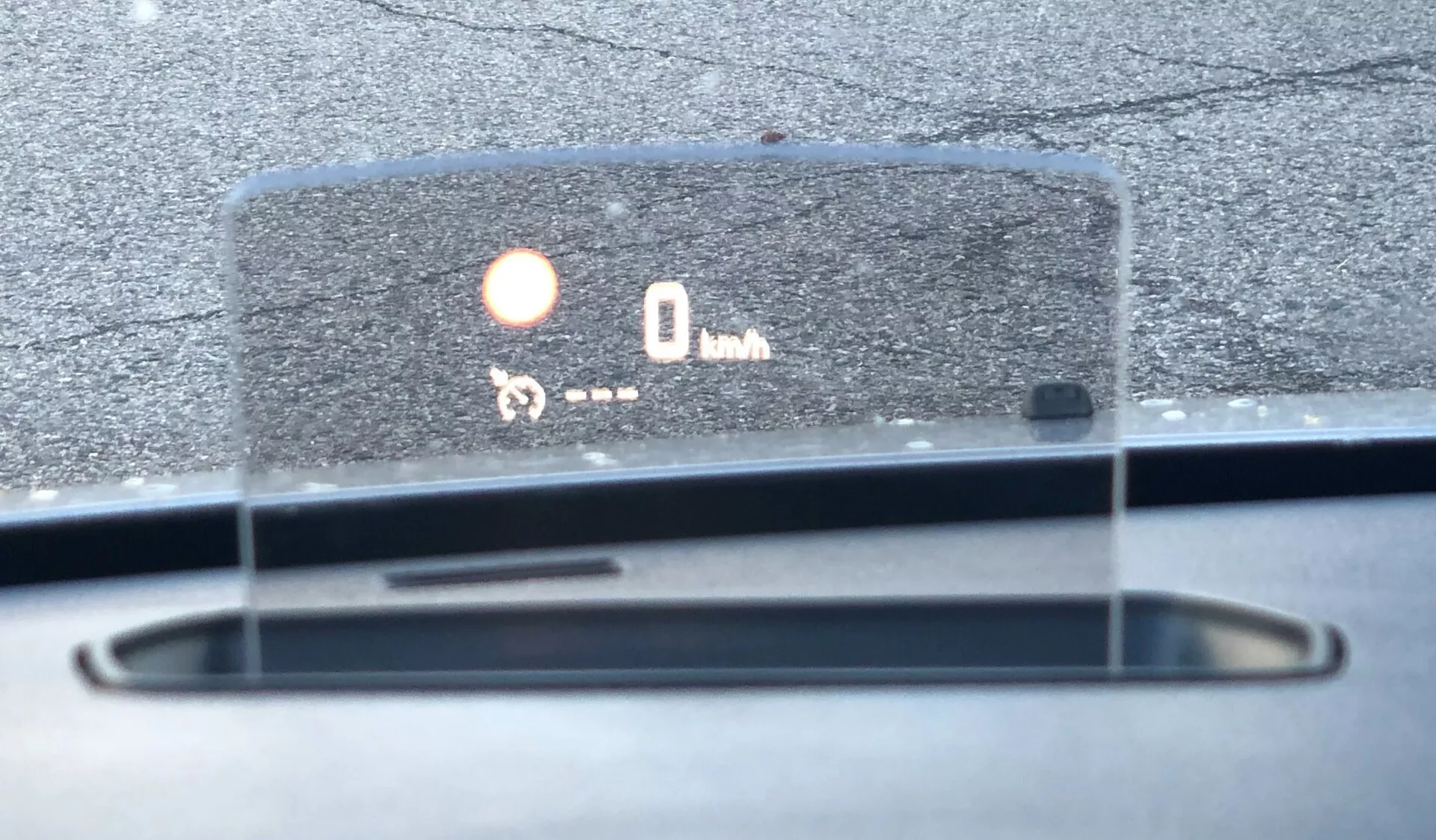 Närbild av head-up display (HUD) i Citroen Spacetourer 2016 som projicerar information på vindrutan, visar hastighet (0 km/h) och en ikon, möjligen indikerande parkeringssensor eller kollisionsvarningssystem. Detaljer av instrumentbrädan, avfrostningsventiler, solljuset speglar daginställning utomhus. Vinkeln visar förarens perspektiv, med fokus på HUD.