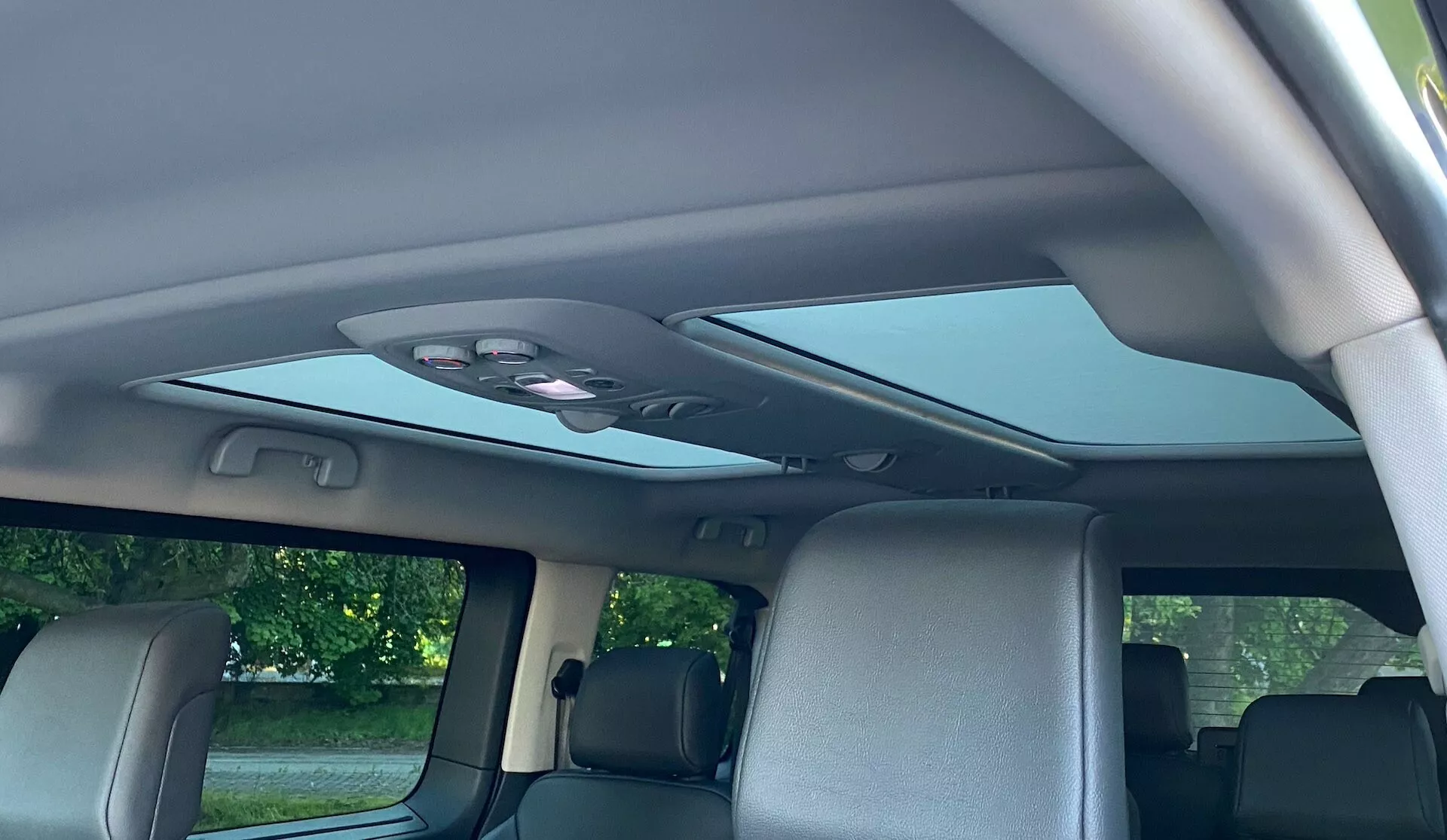 Interiör av en Citroen Spacetourer 2016 sedd från passagerarsätets perspektiv, med ett stort panoramasoltak, delvis indraget täckpanel, överliggande konsol med kontrollknappar och kupollampa, delvis synlig andra radens säten med nackstöd, handtag ovanför de bakre dörrarna, ytterligare inre läslampor, övre monteringpunkter för de bakre säkerhetsbältena och fönster som indikerar ett högt takfordon som en SUV eller van.