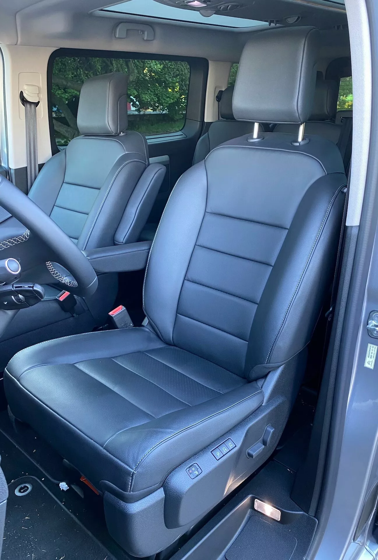 Interiörvy av en 2016 Citroen Spacetourer som framhäver framsätena. Bilden, tagen från sidodörren, visar svarta lädersäten med nackstöd och sidobolster, vilket antyder komfort och stöd. Till vänster syns ratten och en del av instrumentpanelen, med heltäckningsmatta på golvet och en skymt av baksätet, allt under naturligt dagsljus, vilket betonar en rymlig och välskött bil.