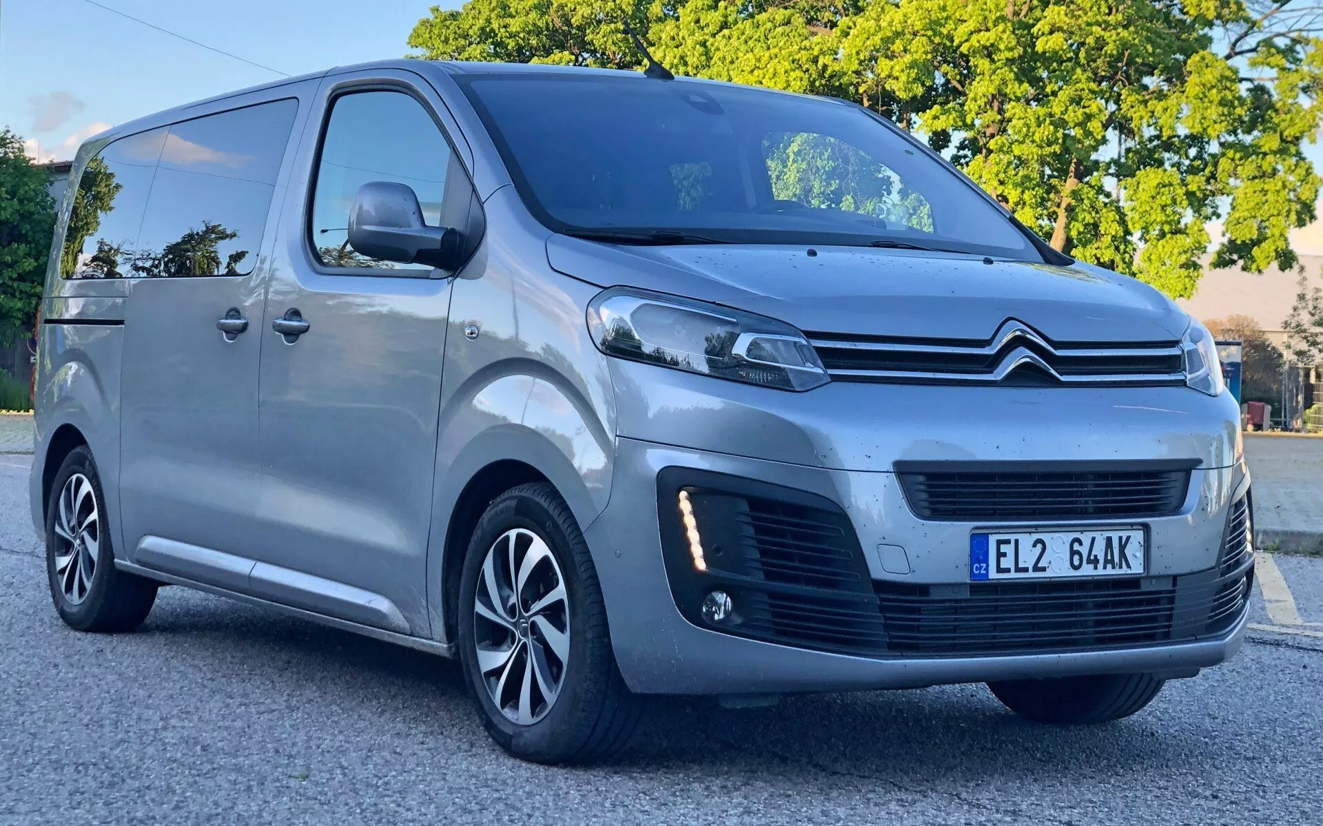 Bild av en Citroën Spacetourer 2016 sedd från en trekvarts främre vinkel, som visar frontgrillen med en kromad Citroën dubbelchevronlogotyp, eleganta strålkastare som sannolikt innehåller LED-dagljus och dimljus under. Skåpbilen har stora sidorutor, lättmetallfälgar och en skjutdörr på sidan, vilket indikerar en design både för passagerare och lätt kommersiellt bruk. Den är parkerad på en gata i dagsljus, vilket ger en tydlig vy över dess tekniska funktioner och övergripande design.
