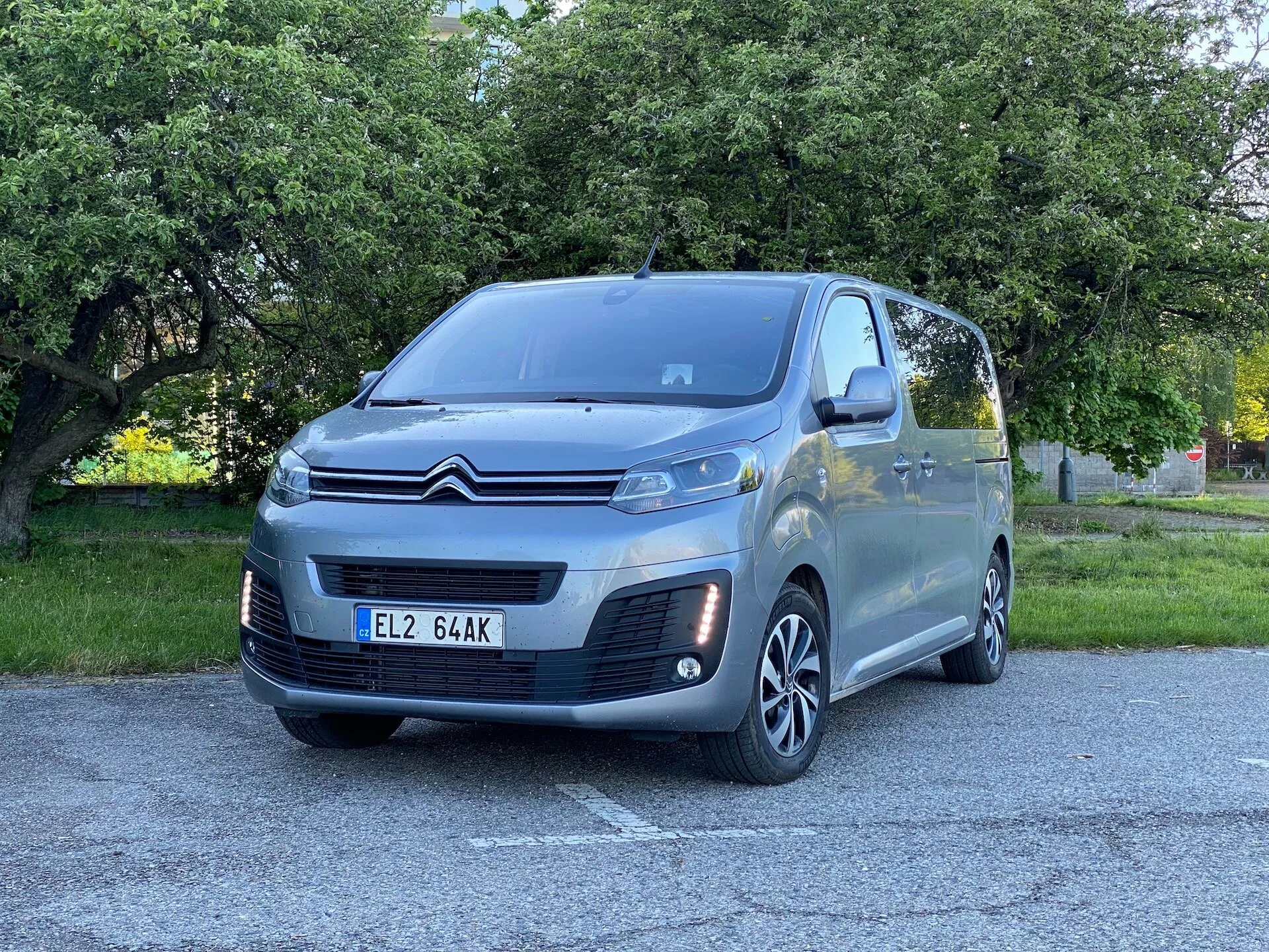 Bilden visar en grå Citroën Spacetourer 2016 passagerarminivan sett från en trekvarts vinkel mot fronten på vänster sida av fordonet. Bilen är parkerad utomhus på ett asfalterat område med grönska, troligen en stadspark eller en trädbevuxen gata. Synliga nyckelinslag är: Citroën-logotyp i form av ett chevron på den svarta och kromade frontgrillen, strålkastardesign med moderna lamphus, dimljus med kromkant, flerspoked lättmetallfälgar, karossfärgade ytterbackspeglar med integrerade blinkers, stora fönster med toning på andra raden, högt taktyp typiskt för en minivan och skjutdörrsspår för baksätespassageraråtkomst. Fotot fångar dessa detaljer från en låg till medelhög vinkel nära bilen, vilket betonar karossens textur och hjulens stil.