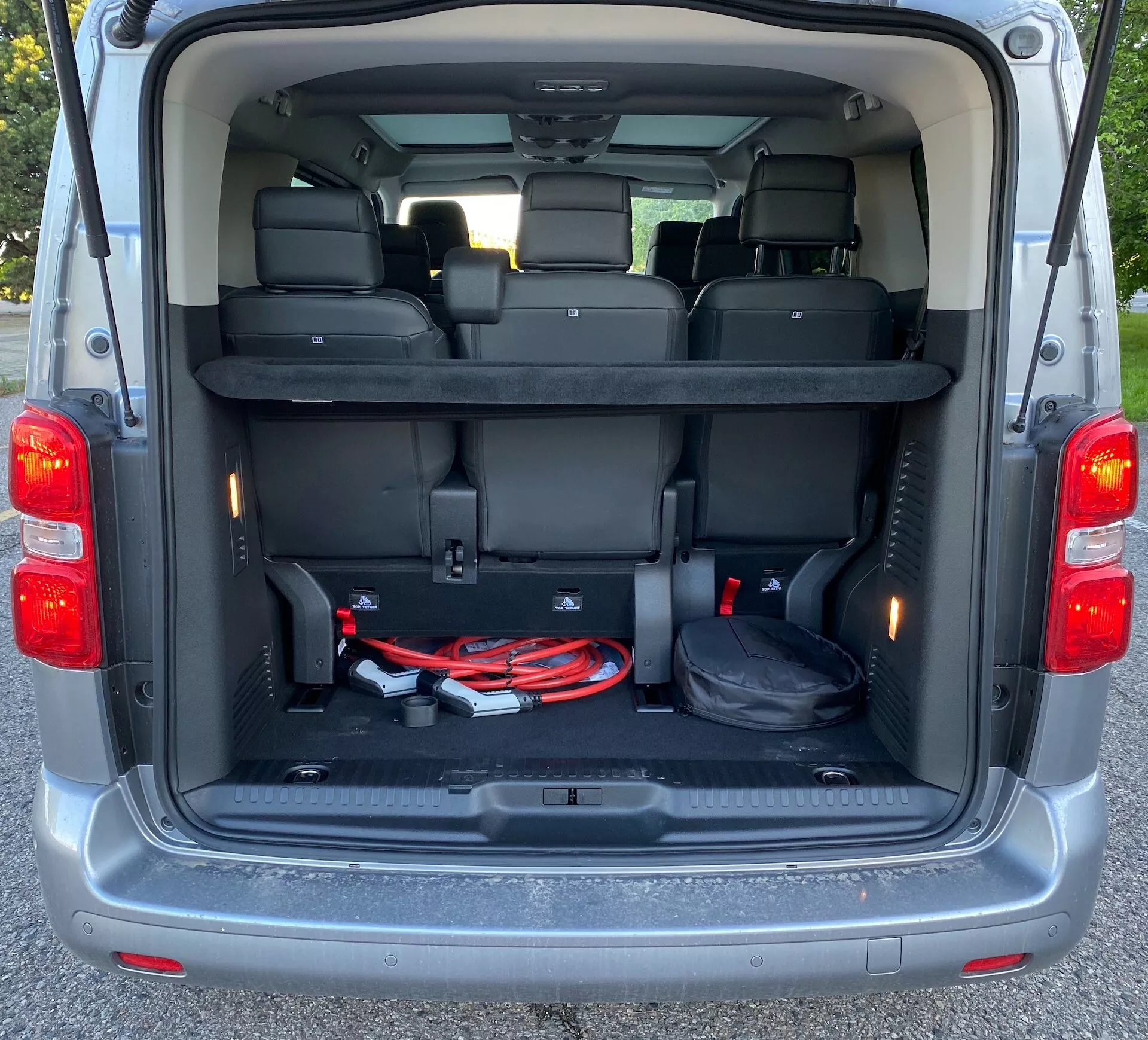 Bakre interiörvy av en Citroen SpaceTourer 2016, som visar det rymliga lastutrymmet med uppfälld baklucka. Tredje sätesraden är i upprätt läge, och den rena och välskötta mattan är synlig. Lastutrymmet har plastlister med förvaringsfack, innerbelysning på sidorna, ett öppet fack på vänster sida och en fördjupning på höger sida för ytterligare förvaring. En startkabel och en svart skyddsmatta syns också på golvet.