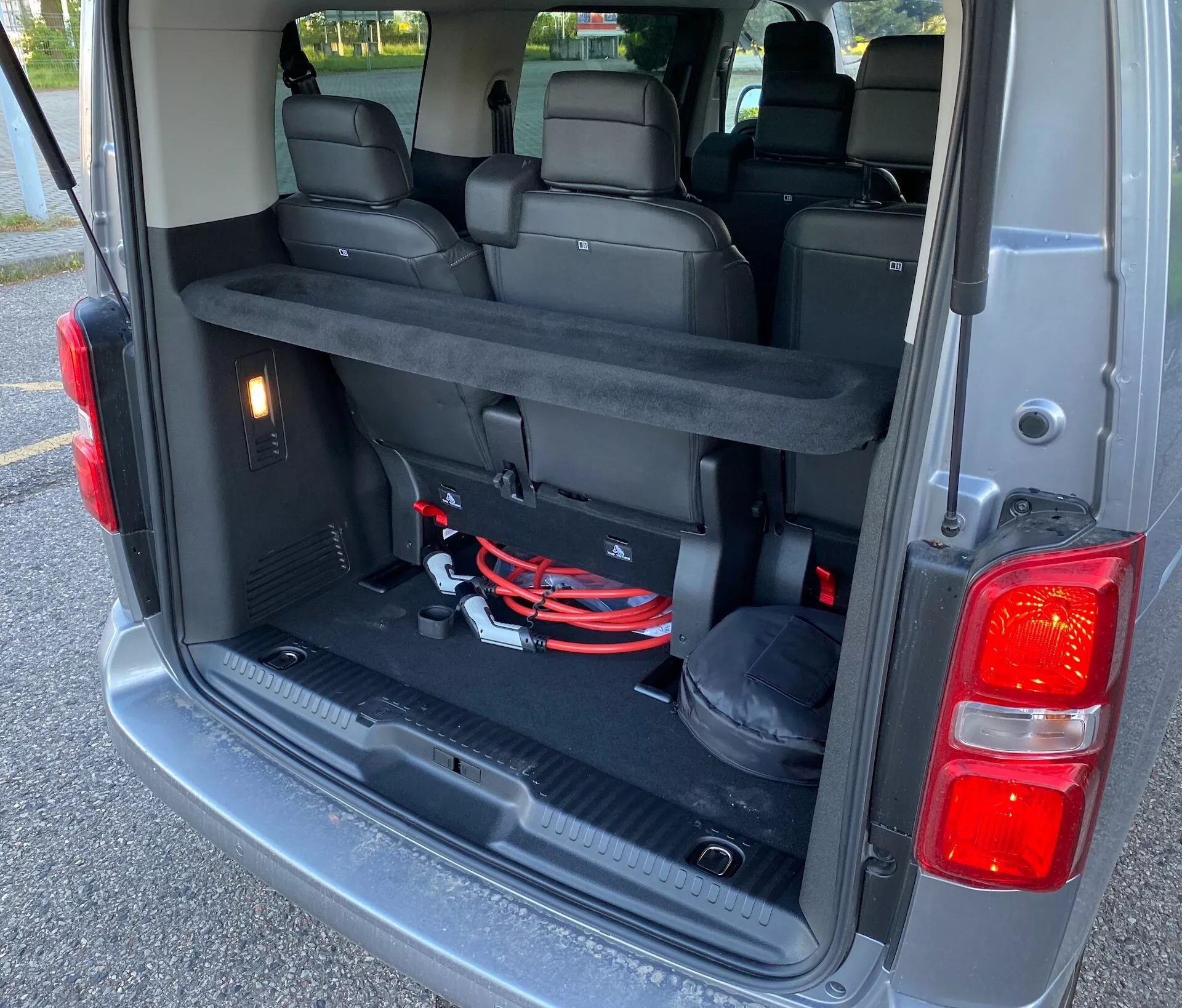 Baksätesvy på en Citroen Spacetourer 2016 från en vinkel bakifrån som visar lastutrymmet med den bakre luckan öppen. Bilden fokuserar på det rymliga utrymmet med den tredje sätesraden nedfälld, vilket ökar lastutrymmet. Fordonet har plats för 7 eller 8 passagerare med delbara nedfällbara säten för mångsidiga arrangement. Synliga funktioner inkluderar innerbelysning, ett 12V-uttag och synliga föremål som startkablar, vilket tyder på ett välutrustat arbetsfordon. De mörkfärgade inredningsdetaljerna, troligen en blandning av tyg eller läder och plastmaterial, samt designen på den bakre luckan inklusive vertikala baklyktor och bakre stötfångare är också synliga.
