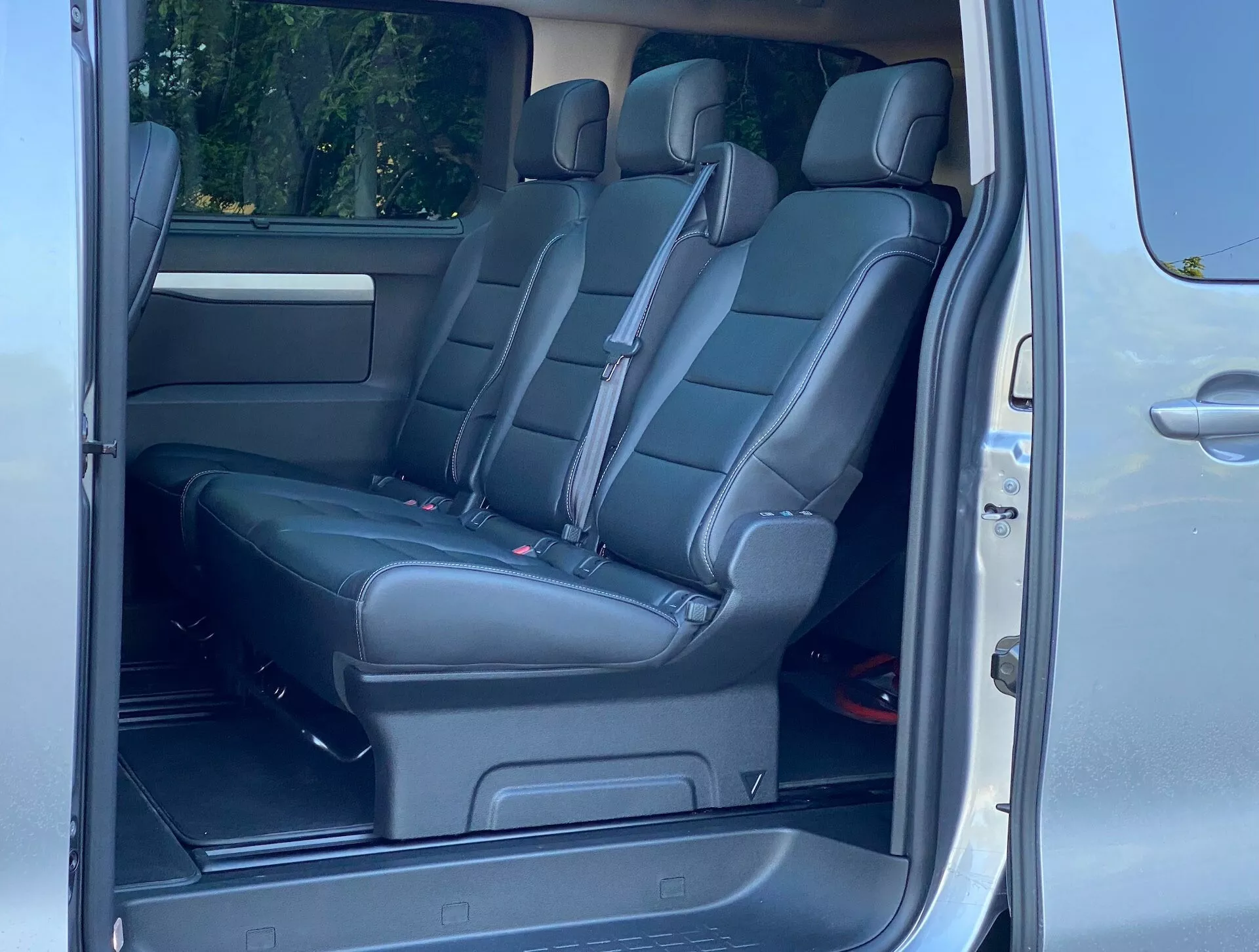 Interiör av Citroën SpaceTourer 2016, fokuserad på passagerarsätena. Bilden är tagen från en sned vinkel vid den bakre passagerardörren, vilket avslöjar andra och tredje raden med säten. Den andra raden består av två individuella svarta lädersäten, medan den tredje raden har en bänk för tre passagerare. Funktioner inkluderar nackstöd, röda säkerhetsbältesknäppningar, uppfällt mittarmstöd, högtalargaller på dörren, klimatanläggningsventil, kopphållare och golvmonterade skenor för justerbara säten.