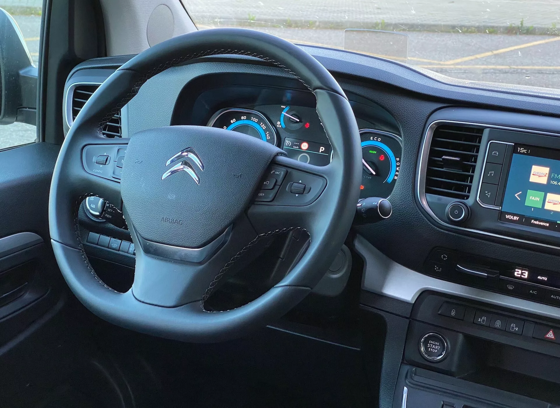 Interiörvy från förarsätet i en modern Citroën Spacetourer 2016 med fokus på ratten och instrumentpanelen. Ratten med inbyggda kontroller antyder integration av ljud, telefonanslutning och farthållare. Det upplysta instrumentklustret inkluderar analoga hastighetsmätare och varvräknare med en central digital display. Infotainment-skärmen som visar en karta, fysiska knappar för media- och fordonskontroller och klimatkontroller indikerar avancerad teknologi och komfortfunktioner. En kontrollpanel med knappar för förarassistanssystem är också synlig. Bilden är tagen från en något förhöjd vinkel, som visar en väl upplyst interiör med naturligt ljus.