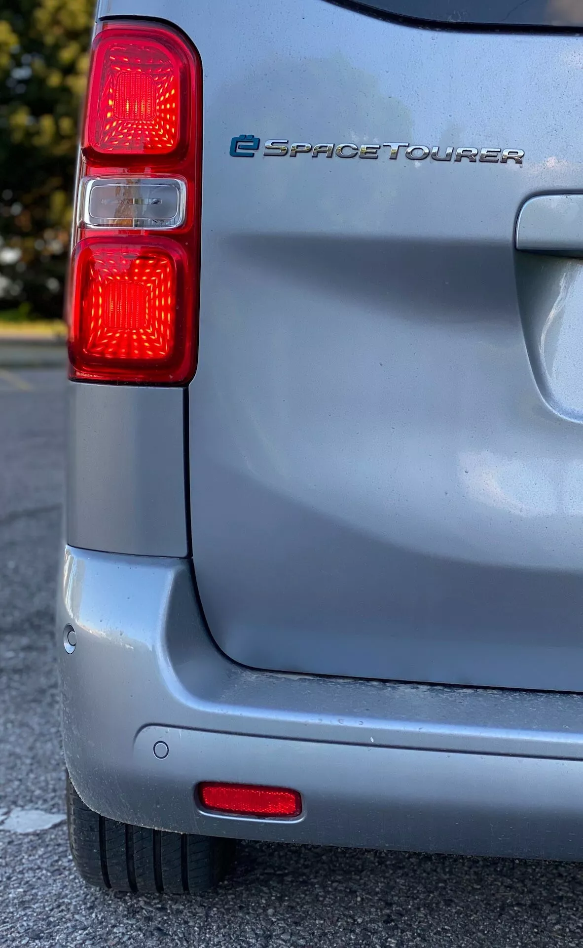 Närbild av bakre delen av en 2016 Citroen e-SpaceTourer elbil tagen från en lågt till mellanhögt perspektiv, som visar den vänstra bakljusgruppen med vertikala röda ljus och integrerade bakre parkeringssensorer i stötfångaren, mot en bakgrund av en solig himmel som framhäver den silverfärgade lacken och den lätta slitage på fordonets yta.