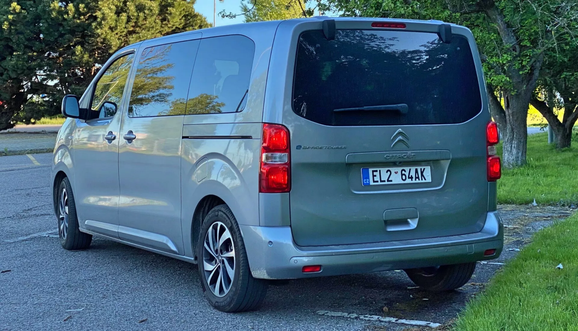 Bakre vy av en metallicgrå 2016 Citroën SpaceTourer van, fångad från en trekvartsvinkel som betonar vänster sida, som visar bakluckan, bakrutan, en tvåfärgad stötfångare med parkeringssensorer och ett upplyst bakre vänster ljuskluster. Bilens skjutdörr med synlig skena ovanför bakhjulet och aluminiumfälgar antyder en blandning av stil och funktionalitet, i en förortsbild som antyder dagsljus, möjligen eftermiddag. Tonat privatfönster och den övergripande standardkonfigurationen framhäver fordonets lämplighet för personligt, familje- eller småföretagsbruk.