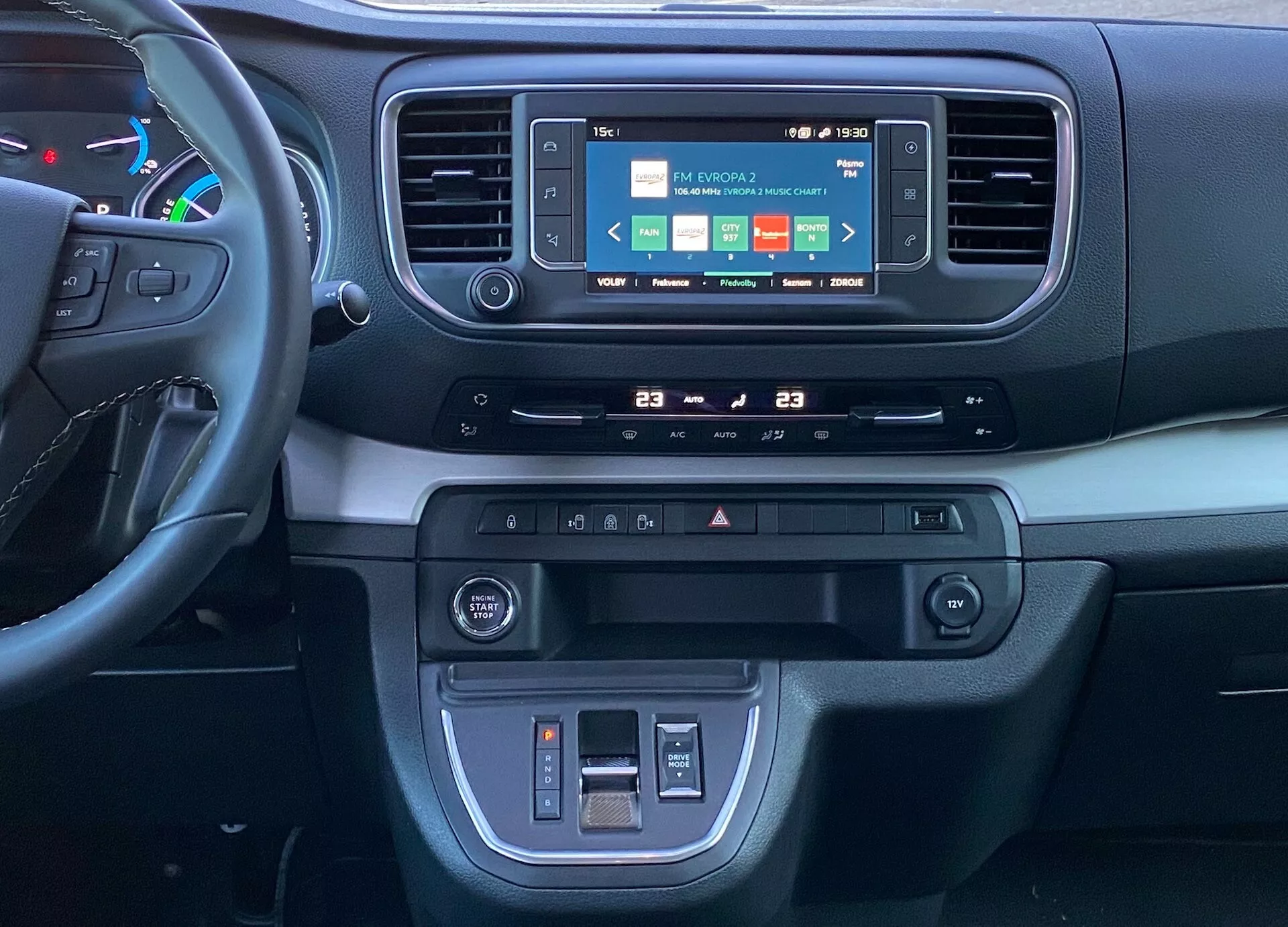 Vy av Citroen Spacetourer 2016 interiör, med fokus på instrumentbrädan och mittkonsolen från framsätespassagerarens perspektiv. Funktionerna inkluderar en färgstark infotainment pekskärm som visar navigation, ljud och telefonanslutning, en automatisk tvåzons klimatanläggning inställd till 23 grader Celsius, knappar för säkerhetsfunktioner inklusive varningsblinkers och förarassistanssystem, 12V-strömuttag och USB-port för laddning och medieanslutning, ett litet förvaringsfack, en kombination av mjuka och robusta plastmaterial, en fragmenterad multifunktionsratt och en ergonomisk layout som följer moderna fordon designprinciper.