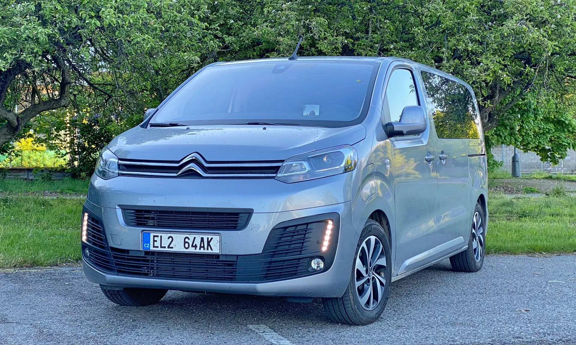 Bilden visar en modern Citroen SpaceTourer 2016 placerad diagonalt, med fronten lite åt höger. Förarsidan, hela fronten och delvis sidoprofil är synliga. Bilen upptar en stor del av ramen, med betoning på sina LED-strålkastare, distinkta främre grill, flerspåriga lättmetallfälgar, sidospeglar med indikatorer och parkeringssensorer på främre stötfångaren. Den metalliska finishen och eleganta designen betonar modern fordonsingenjörskonst. Scenen utspelar sig utomhus i en parklik miljö under dagtid, kännetecknad av naturligt ljus och bakgrundsträd och gräs.