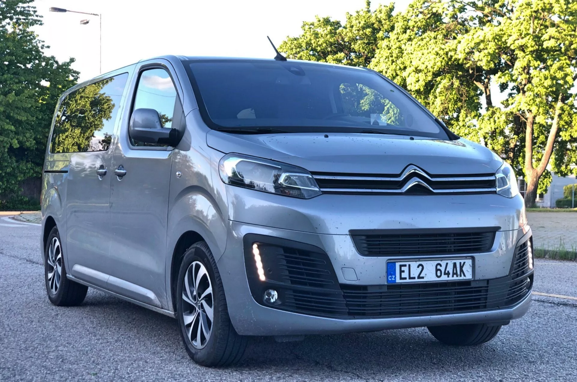 Låg vinkel, vy i trekvartsfront av en metallicgrå Citroën Spacetourer MPV 2016-modell, som framhäver den framstående frontgrillen med dubbelchevron-logotyp, skulpterade LED eller HID-strålkastare, parkeringssensorer i stötfångaren, flerspakiga aluminiumfälgar, strömlinjeformade sidospeglar med integrerade blinkers och stora fönster med stigande fönsterlinje. Bilen står parkerad på en asfaltsyta i en förorts- eller parkliknande miljö med lövverk i bakgrunden.