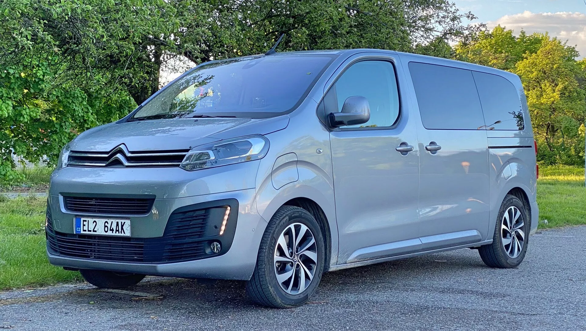 Ett foto av en Citroën Spacetourer 2016 skåpbil taget från en låg framvinklad trekvartsvy, som visar bilens fram- och sidoprofil. Det eleganta, moderna designen inkluderar en framträdande Citroën dubbel chevron-logga på grillen, distinkta strålkastare möjligen med LED-dagljus, en centrerad främre registreringsskylt och ett nedre luftintag på främre stötfångaren. Fordonet är målat i ljus silvergrå färg med stora sidospeglar och flerspoke lättmetallfälgar, vilket antyder en medel- eller högre utrustningsnivå. Sidoprofilen inkluderar en skjutdörr på förarsidan och solida panelsektioner för lasttransport. Bakgrunden består av vägkantens grönska, fångad i naturligt dagsljus.