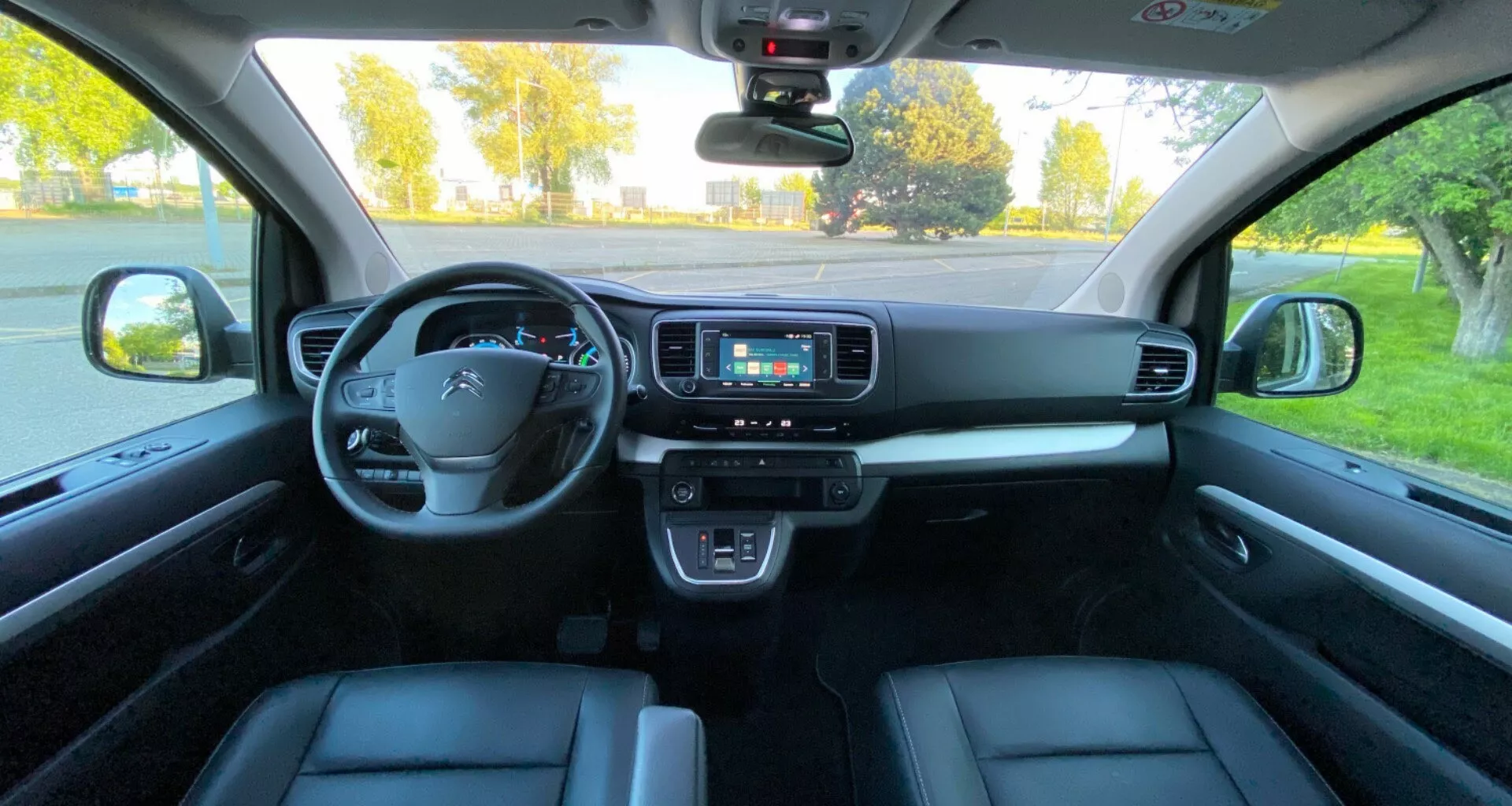Interiörvy av Citroen Spacetourer 2016 från baksätets perspektiv, som visar en ratt med integrerade kontroller, digitalt instrumentkluster, mittkonsol med infotainmentskärm, klimatanläggningsknappar, växelspak, elektronisk parkeringsbromsknapp och dörrkontroller. Modern fordons teknik och ergonomi är uppenbar med borstat metall- eller träfinish och koordinerad färgskala.