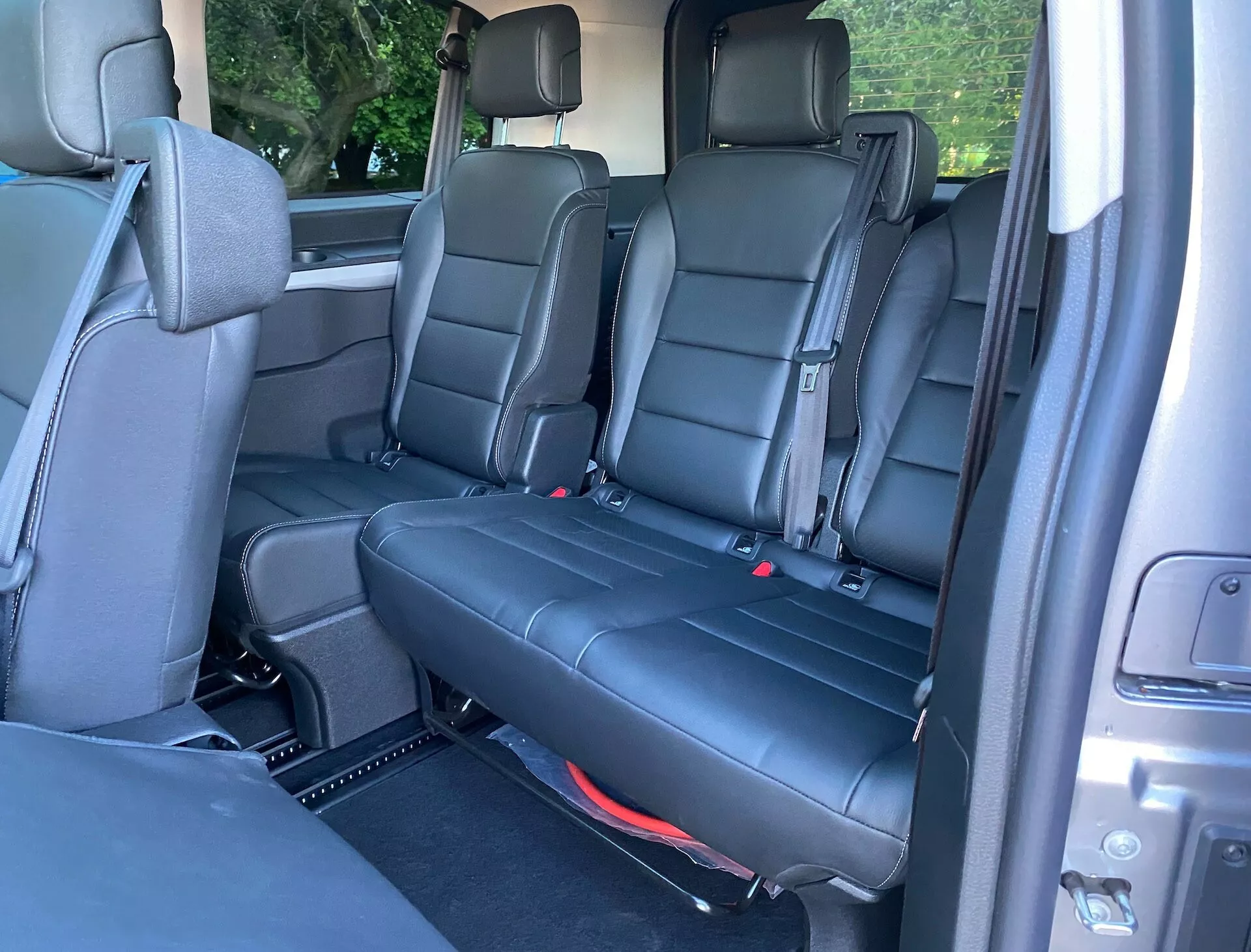 Interiörvy av Citroen Spacetourer 2016 som visar baksätesområdet från en sned vinkel, med fokus på de svarta lädersätena i andra och tredje raden. Andra raden har två separata säten med armstöd och gångavstånd, medan tredje raden har en bänktyp. Synliga element inkluderar säkerhetsbälten, röda barnstolsfästen, kontrollmekanismer, ventilationsuttag, mattbeklädd golv med avtagbara mattor, högtalare och elektriska fönsterkontroller. Naturligt ljus framhäver en design inriktad på familjekomfort.