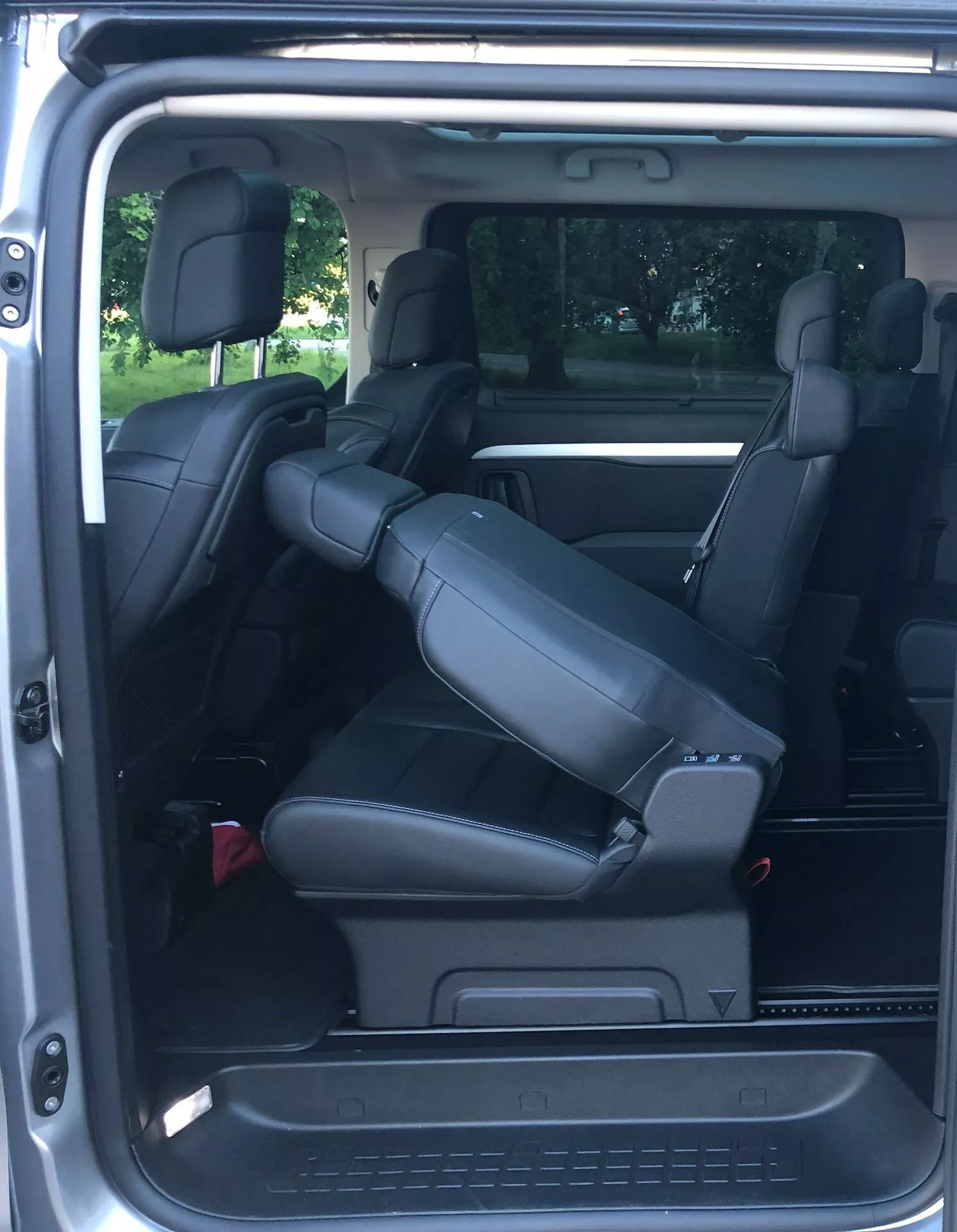 Interiörvy av en Citroën Spacetourer 2016 från bakifrån, med fokus på baksätena. Den moderna minibussen har tre rader med säten. Den mittersta raden, med kaptensstolar, är lutad framåt vilket visar att de går att fälla för att öka lastutrymmet eller komma åt tredje raden. Den bakre raden har också två säten med nackstöd och gott om benutrymme. Interiören är klädd i svart läder för en premiumkänsla, med säkerhetsbälten och en klimatanläggningsventil på sidopelaren som visar bakre klimatanläggningsfunktioner.
