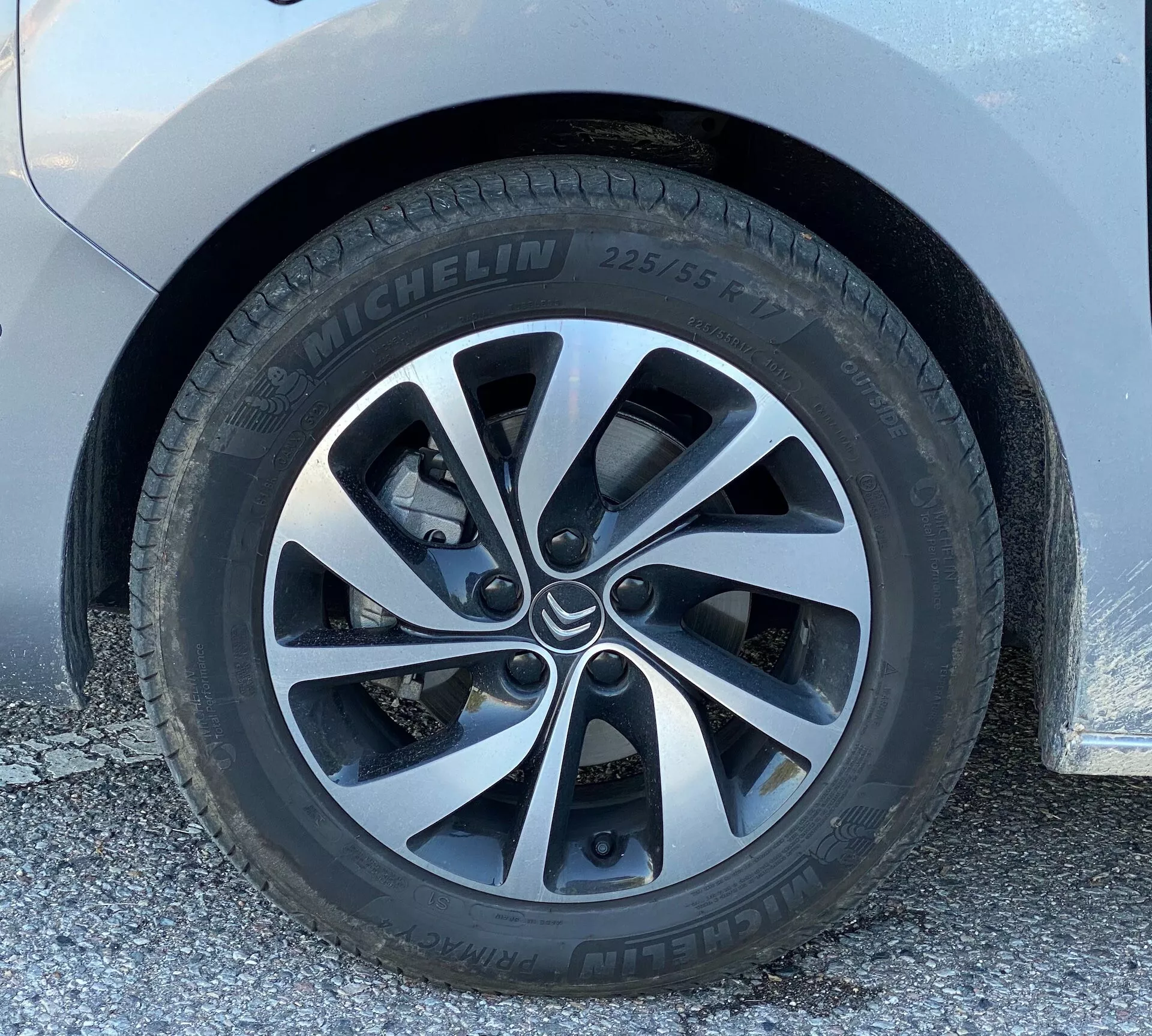 Närbild av 2016 Citroen Spacetourer hjul från låg vinkel, nästan i marknivå, framhäver en Michelin-däck (225/55) och ett modernt legeringshjul med fem parade ekrar. Hjulet är rent, med minimalt smuts, som visar en del av skivbromssystemet genom ekrarna. Karossen ovanför hjulet är ljusfärgad, möjligen vit eller silver.