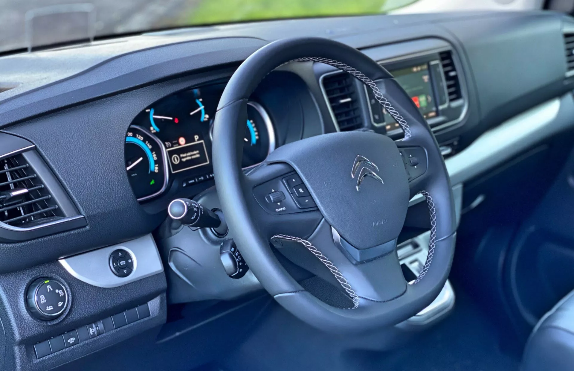 Interiörvy av en Citroen Spacetourer 2016 som fokuserar på förarplatsen, visar ratten med monterade kontroller, instrumentpanelen med analoga mätare och digital display, samt delvis synlig central display och knappar för klimatanläggning på mittkonsolen. Även synliga är fönsterkontrollerna och sidobackspegeln med elektriska justeringar på förardörren.